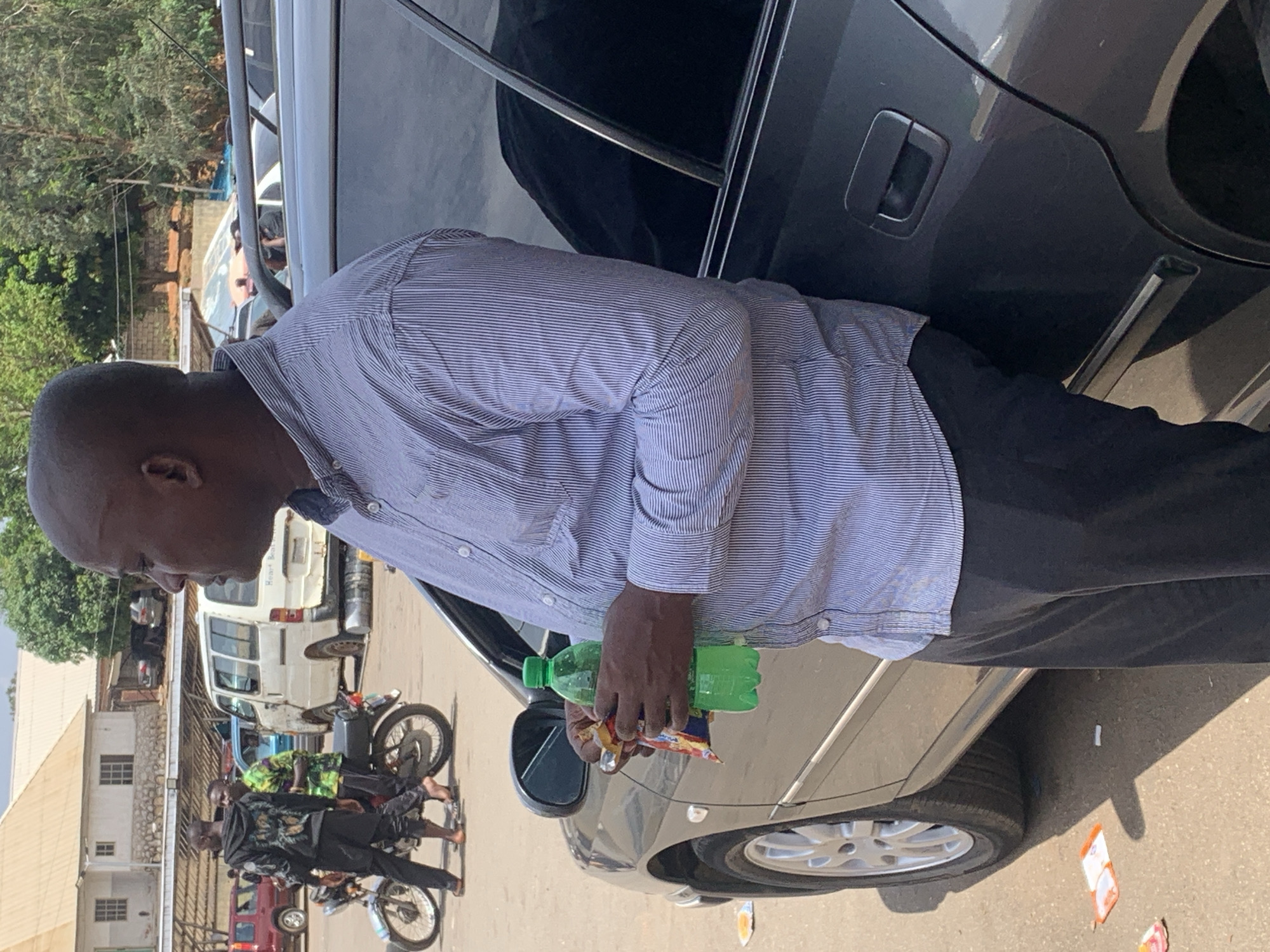 A middle-aged, Black man stands beside a dark gray car on a street in Bukuru, Nigeria. He wears a light blue and white striped button-down shirt and dark pants. In his hand, he holds a green plastic bottle and a package of snacks. The car is parked on the side of the street, and other cars and motorcycles are visible in the background. Buildings and trees line the street. The scene suggests a casual, everyday moment in a busy urban setting.