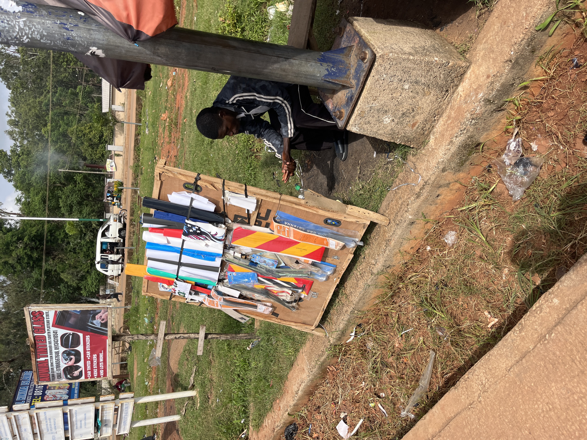 In Bukuru, Nigeria, a street vendor sits near a display of automotive accessories. The vendor, a young Black man wearing a dark jacket with white stripes and dark pants, is seated on a low bench, his hands clasped in front of him. He is positioned to the right of a wooden display board that showcases various car accessories, including car tinting films in multiple colors, car stickers, and what appear to be reflective safety strips. The display board is propped up on a wooden stand.
The environment is a roadside setting. To the left of the vendor, a concrete drainage ditch runs along the edge of the road, filled with some debris and dry grass. Beyond the ditch, there is a grassy area and several signs on poles. One prominent sign advertises "AOWAL TINTED GLASS" and lists services like "CAR TINTED," "CAR STICKERS," and "AND LOTS MORE..." along with a phone number. In the background, a road is visible with a white van and a tricycle passing by. Lush green trees form a backdrop to the scene. The overall impression is one of an outdoor market or roadside stall in a moderately busy urban area.