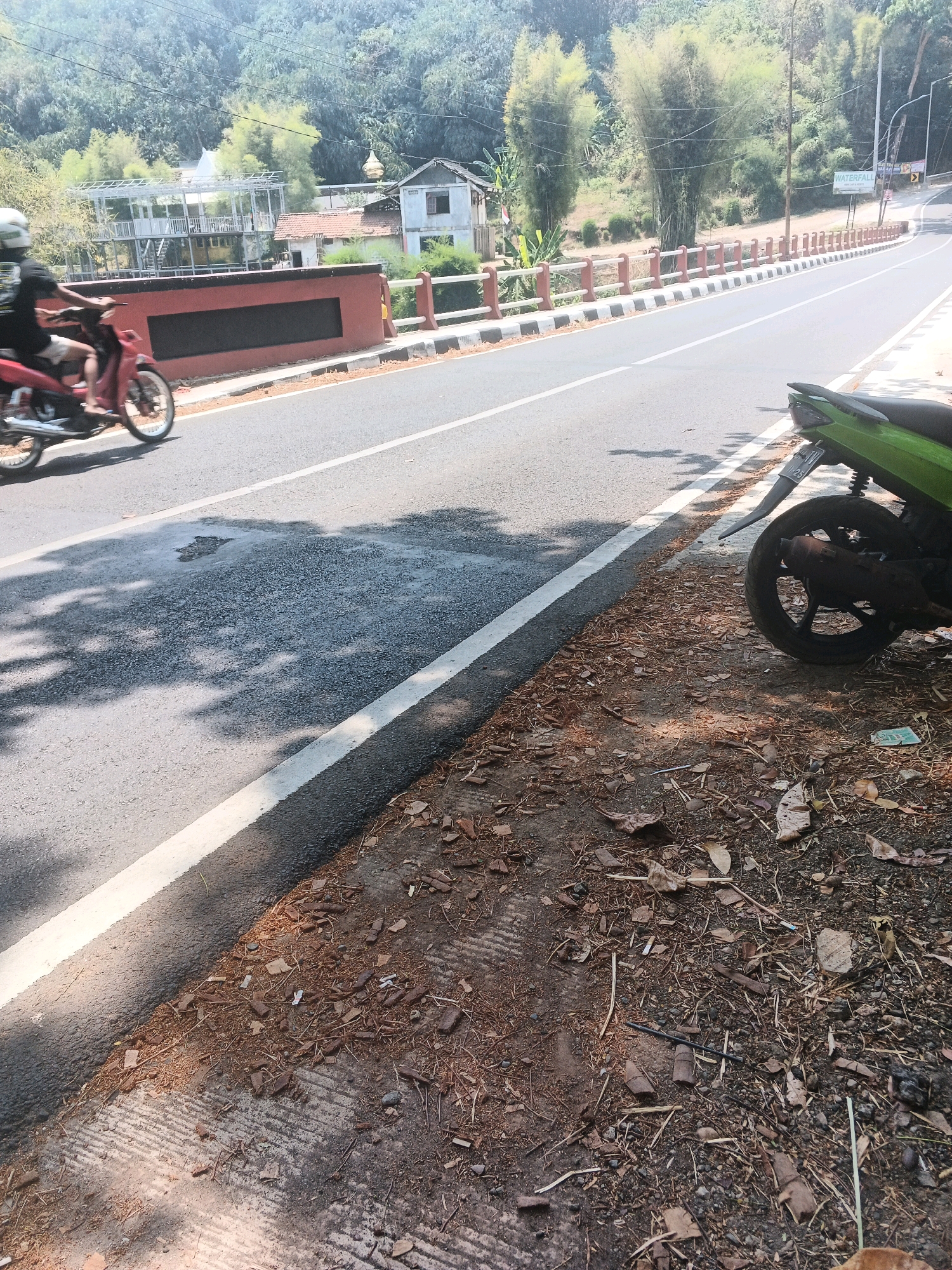 A paved road runs diagonally from the lower left to the upper right of the image. On the left side of the road, a person is riding a red motorcycle. The motorcycle's wheels are blurred due to motion. A white line marks the edge of the road on the left. The road surface appears wet in places, with shadows of trees cast upon it. On the right side of the road, a green motorcycle is parked. The area to the right of the road is covered in dry leaves, dirt, and gravel. In the background, there are trees, a building with a red roof, a white structure with scaffolding, and a bridge with red and white railings. A sign with the word "WATERFALL" is visible in the distance. The overall setting appears to be a rural or semi-rural area outside of a city. The presence of a motorcycle and a paved road suggests it is accessible by vehicle. The vegetation and natural elements indicate an outdoor environment, likely in a tropical climate due to the lush greenery. The presence of a sign pointing to a waterfall suggests it could be a tourist destination or a natural attraction. Based on the architecture and vegetation, the location is consistent with Southeast Asia, specifically Indonesia.
