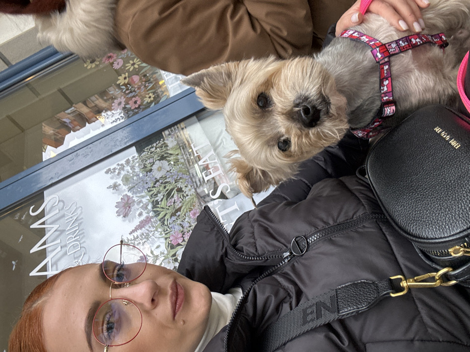 This image depicts two individuals and a dog situated outside an establishment identified as "AMS & DRINKS" and "LET'S EAT" in Budva, Montenegro. The primary subject, a female, occupies the left side of the frame, looking towards the camera. She presents with reddish-orange hair pulled back and wears red-rimmed circular eyeglasses. Her attire consists of a black puffer jacket over a white turtleneck. A black textured shoulder bag with a gold clasp and a visible "FENDI" strap is slung across her body. A mole is observed on her left cheek. Her left hand rests on a small, light brown and gray Yorkshire Terrier-type dog, which wears a pink and red patterned harness and also faces the camera. A second individual, wearing a brown coat with a fur-lined collar, is partially visible on the far right. The background features a large window adorned with multi-colored floral and butterfly decals. A brick building is reflected in the upper portion of the window glass.