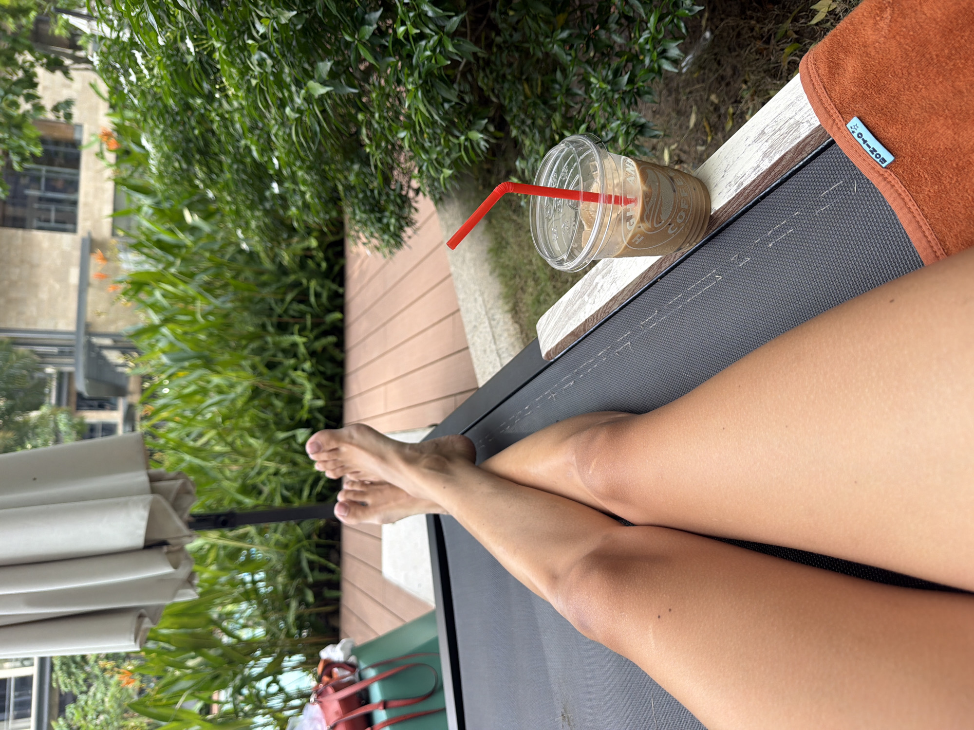 A person's legs are extended on a lounge chair outdoors, with a clear plastic cup of iced coffee with a red straw to the right. The person's feet are crossed at the ankles, with toes pointed upwards. In the background, a white umbrella stands over a wooden deck. Lush green plants and trees are visible behind the deck, with a multi-story building in the distance. An orange towel is draped over the armrest of the lounge chair.