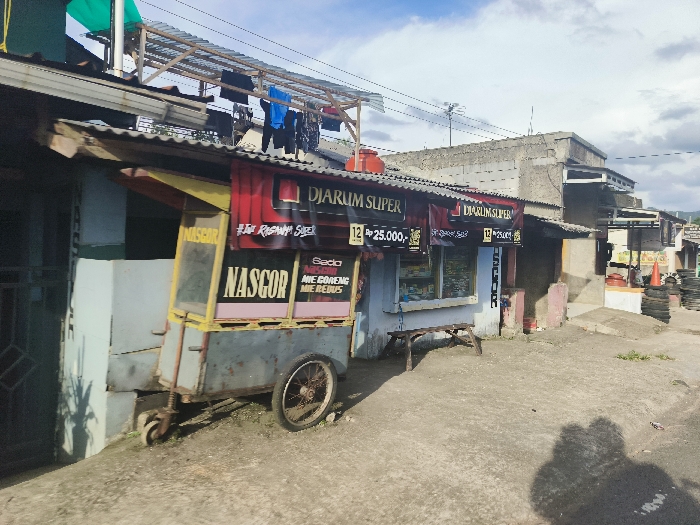 The image shows a street scene in Cikalong, Indonesia, at the coordinates -6.758 latitude and 107.446 longitude. A food cart, labelled "NASGOR" and "Sedo Ane Goreng Meredus," is parked on the left side of the street, facing the viewer. It is a metal cart with a yellow and red canopy and a large metal frame. The cart is in front of a building with a sign that says "DJARUM SUPER." There is a blue plastic bucket in front of the building, which is likely used for collecting water.
The building on the right side of the street appears to be a shop. The sign above the shop is in Indonesian and is not fully visible in the image. There is a wooden bench in front of the shop. The street is paved with concrete and there are some scattered weeds. The sky is clear with some clouds.