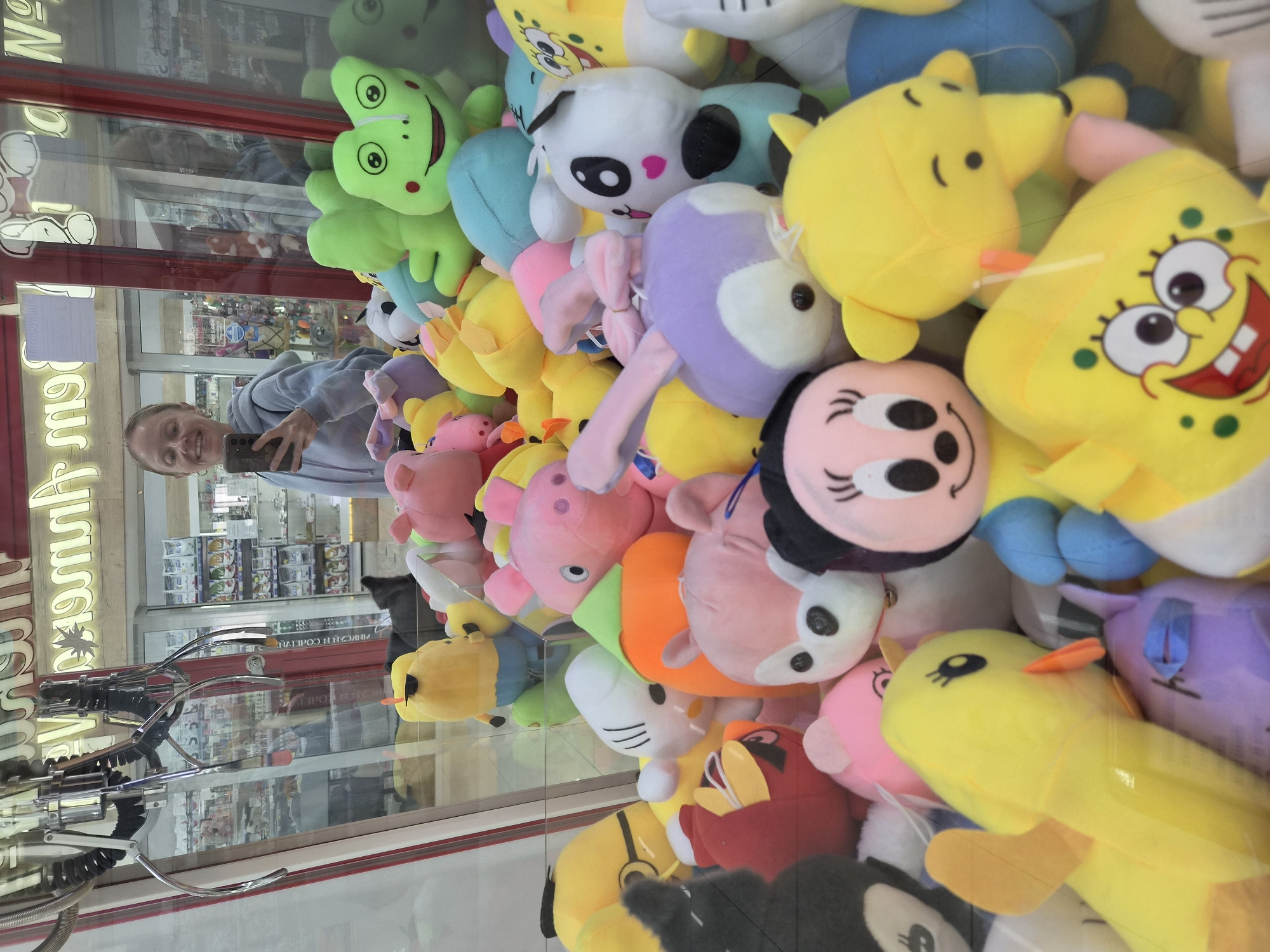 The image shows a claw machine filled with numerous plush toys, located in a commercial establishment in Vladivostok, Russia. A young woman with blonde hair is visible in the reflection of the glass, holding a smartphone and taking a selfie. She is wearing a light blue hooded sweatshirt. The claw machine contains a variety of colorful toys, including a large green frog, a yellow character resembling Minion from Despicable Me, a pink pig, a white character with black ears and a pink heart on its chest, and a yellow Spongebob Squarepants. The background shows shelves with various products, possibly in a store or arcade. The lighting appears to be artificial, typical of an indoor commercial setting.
