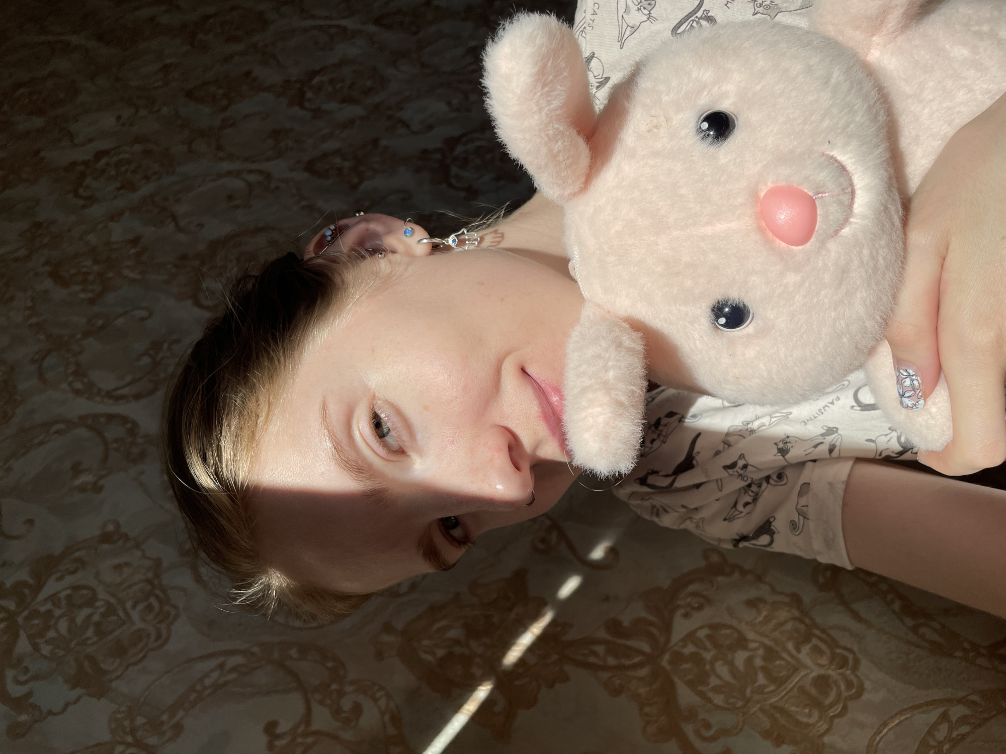 A person is lying on a bed hugging a plush bunny. The person has fair skin and light hair with multiple ear piercings. They have a nose ring. Their nails are painted with a floral design. They are wearing pajamas with a cat pattern. The bed has a patterned beige and brown bedspread. Sunlight is streaming into the room, casting shadows across the bed. The image does not provide information to determine the location of the city of Izhevsk, Russia.