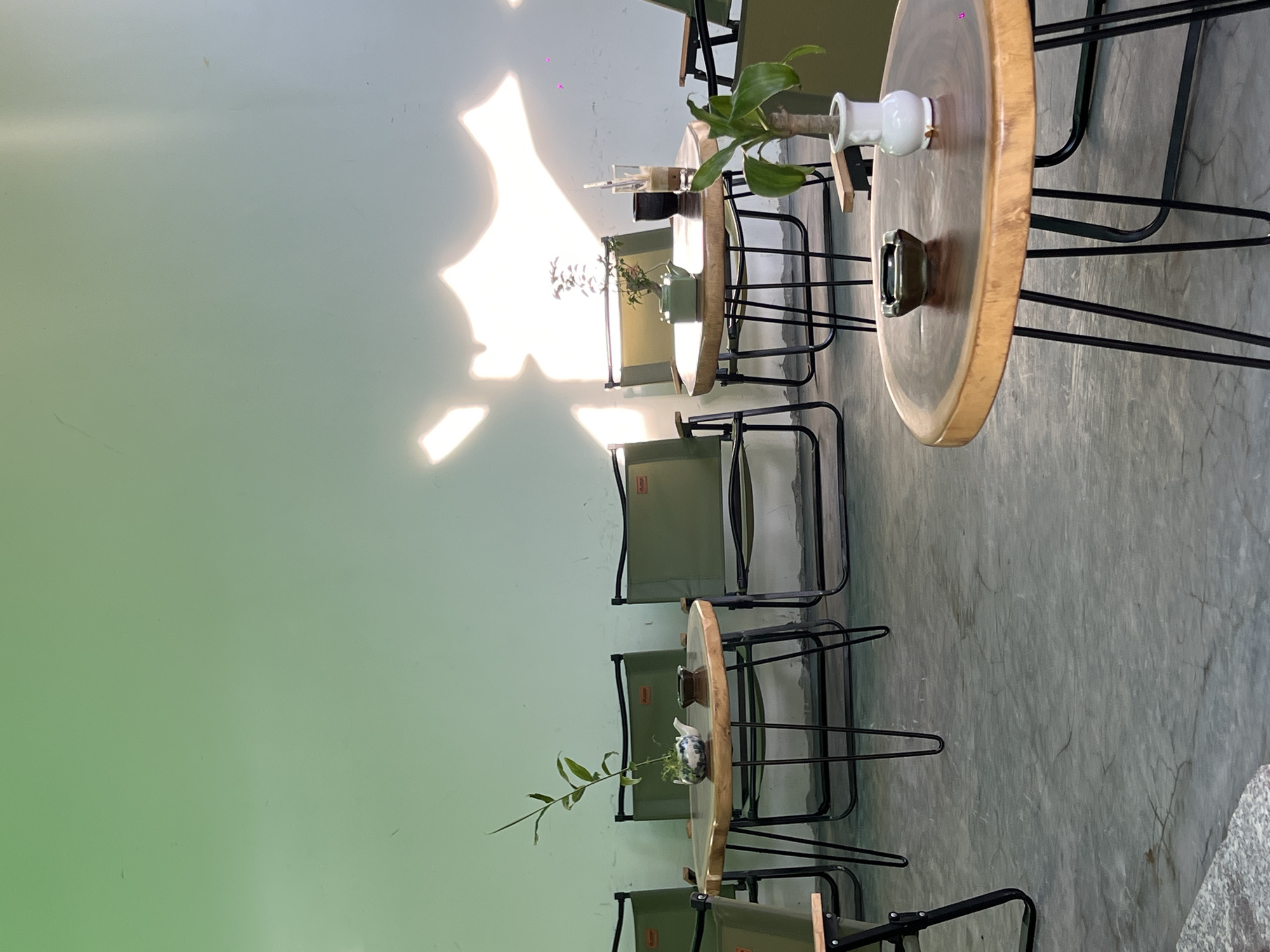 This is an outdoor seating area at a cafe in Nha Trang Ward, Vietnam. The scene is set against a light green wall with sunlight casting a dappled pattern. Several green canvas camping chairs are arranged around round, wooden tables with black metal hairpin legs. One table features a small potted plant and a white teapot with blue floral patterns. Another table has a glass with a straw, a dark cup, and a small green teapot. A third table in the foreground has an ashtray and a white vase containing a leafy plant. The floor is concrete. No people are visible in the image.