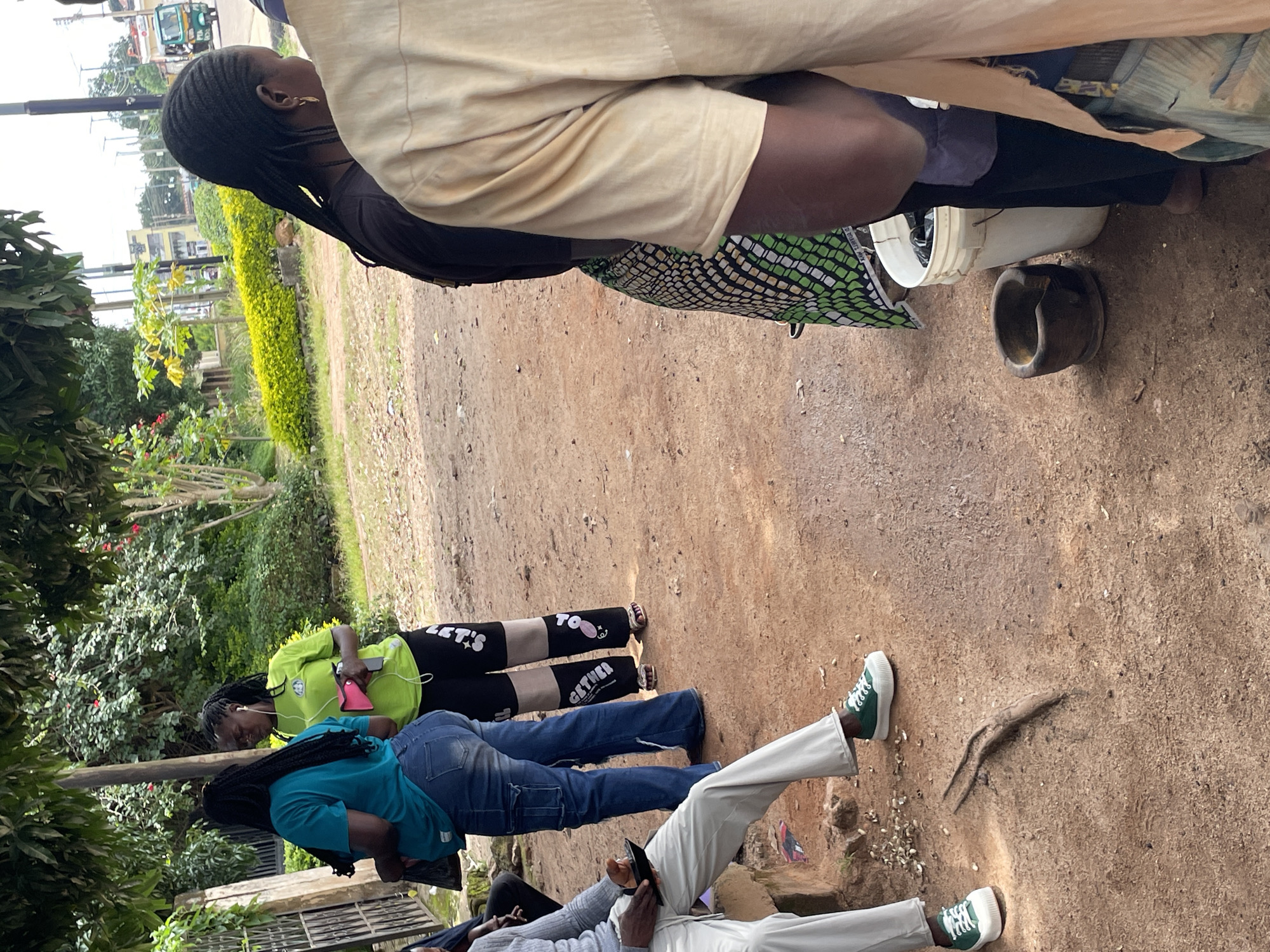 The image depicts an outdoor scene in Jos, Nigeria, likely a market or roadside stall. In the foreground, a person in a light-colored shirt is partially visible, standing next to a white bucket and a wooden mortar. Further back, two women are engaged in conversation. One woman, wearing a green t-shirt and black pants, holds a pink object, possibly a phone. The other woman, in a blue t-shirt and denim jeans, appears to be listening. A man in light-colored pants and green sneakers is seated on a ledge on the left side of the frame, holding a black object, likely a phone. The ground is covered in reddish-brown earth, with scattered debris. In the background, lush green foliage and trees suggest a tropical or subtropical environment. A street with vehicles, including a trishaw, is visible in the distance, indicating an urban setting. The overall atmosphere appears casual and everyday.