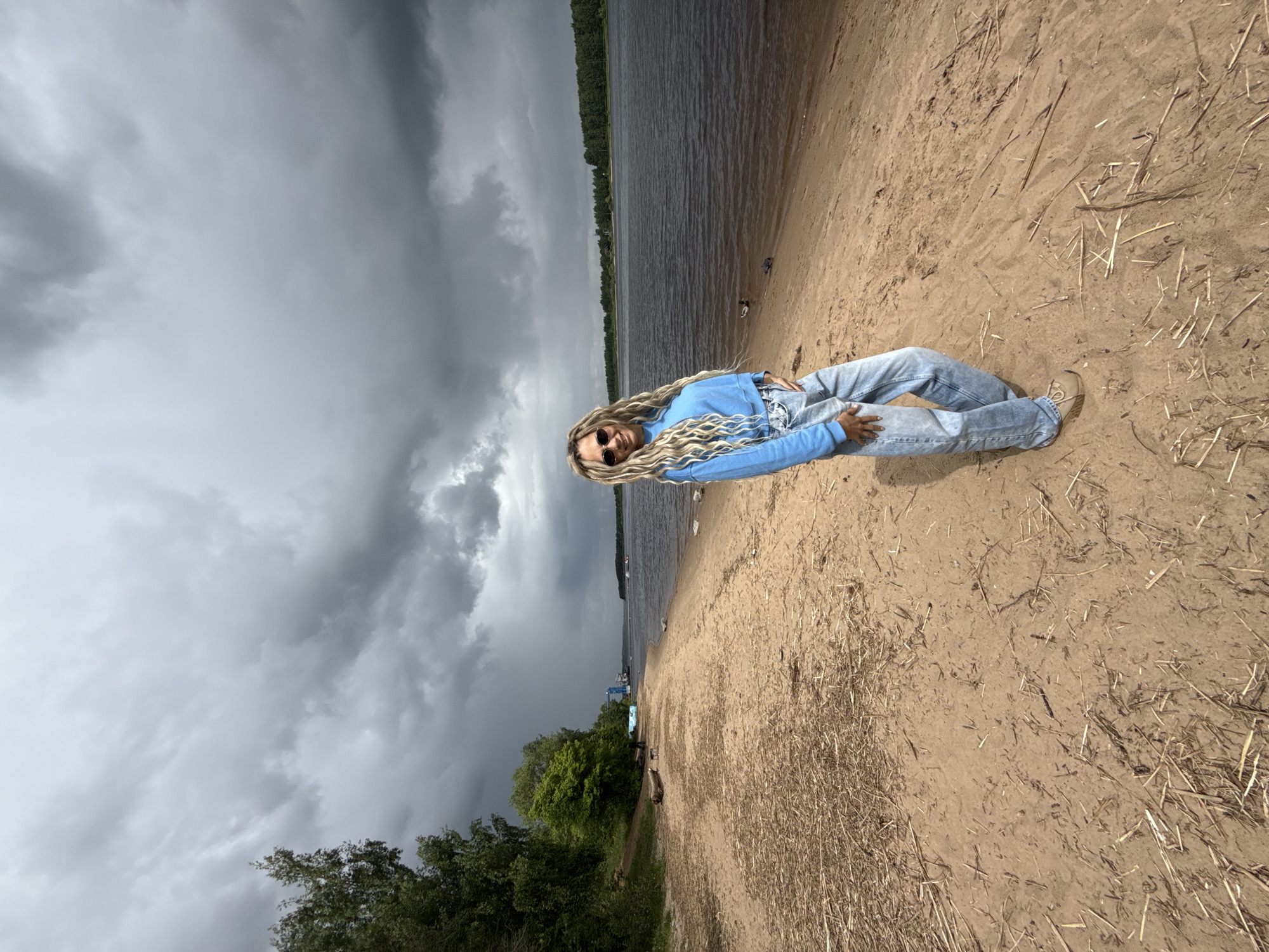 A woman lies on a sandy beach alongside a body of water under a stormy sky. The woman is wearing a light blue sweatshirt, light blue jeans, tan shoes, and sunglasses. She has long, blonde, rope-like braids. The beach is littered with dry grass and twigs. In the distance, there are trees and what appears to be a boat launch. Based on the location in Rusanovo, Russia, the body of water is most likely the Oka River or a nearby lake.