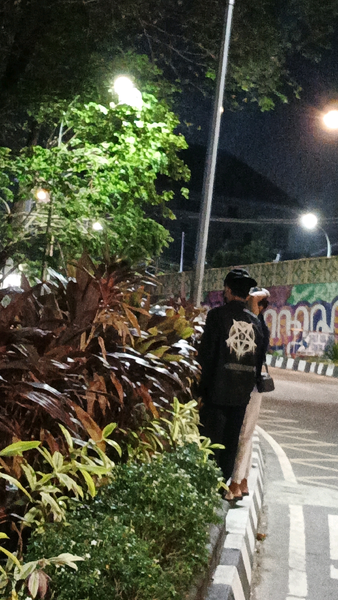 The image depicts a nighttime scene on a street in Surakarta, Indonesia. In the foreground, lush green foliage and plants with reddish-brown leaves line the edge of a sidewalk. A paved road with white markings is visible to the right, bordered by a curb with black and white stripes.
Two individuals are standing on the sidewalk, facing away from the camera and towards the street. The person in the foreground is wearing a black jacket with a prominent white pentagram design on the back. They also appear to be wearing a dark beanie. The second person, partially obscured, is wearing light-colored pants and brown shoes. They are carrying a bag.
In the background, a brightly lit streetlamp illuminates a tree. Further in the distance, a colorful mural adorns a wall, and another streetlamp is visible. The overall atmosphere is nocturnal, with artificial lighting casting shadows and highlighting elements of the urban landscape.