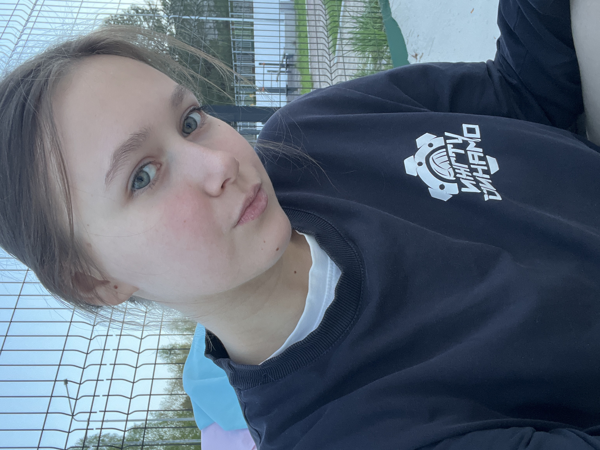 A selfie of a young woman with light skin and light eyes. She is wearing a navy blue sweatshirt with a white logo on the upper right side. She is outside in an urban area near a wire fence and some greenery. Considering the logo on the sweatshirt and the image context, it is likely that the location is Chaikovsky, Russia. The woman is looking at the camera with a slight pursing of the lips.