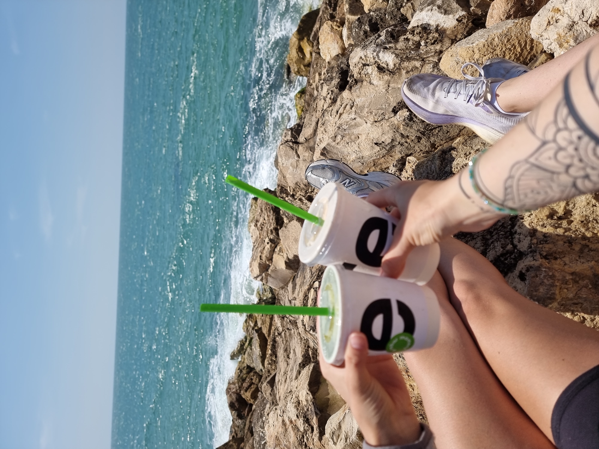 The photograph captures a view of the Mediterranean Sea near Tel-Aviv, Israel, specifically from a rocky cliffside along the coast. The sea is visible to the left, with waves breaking against the rocks below. The sky is clear and blue.
In the foreground, two hands hold cups with green straws, presumably containing drinks. The cups bear a logo. A pair of legs and feet are visible, adorned with sneakers and a tattoo on one arm. The person is seated on the rocky terrain. The focus is split between the refreshments, the coastal view, and the seated individuals, implying a relaxed and social setting.