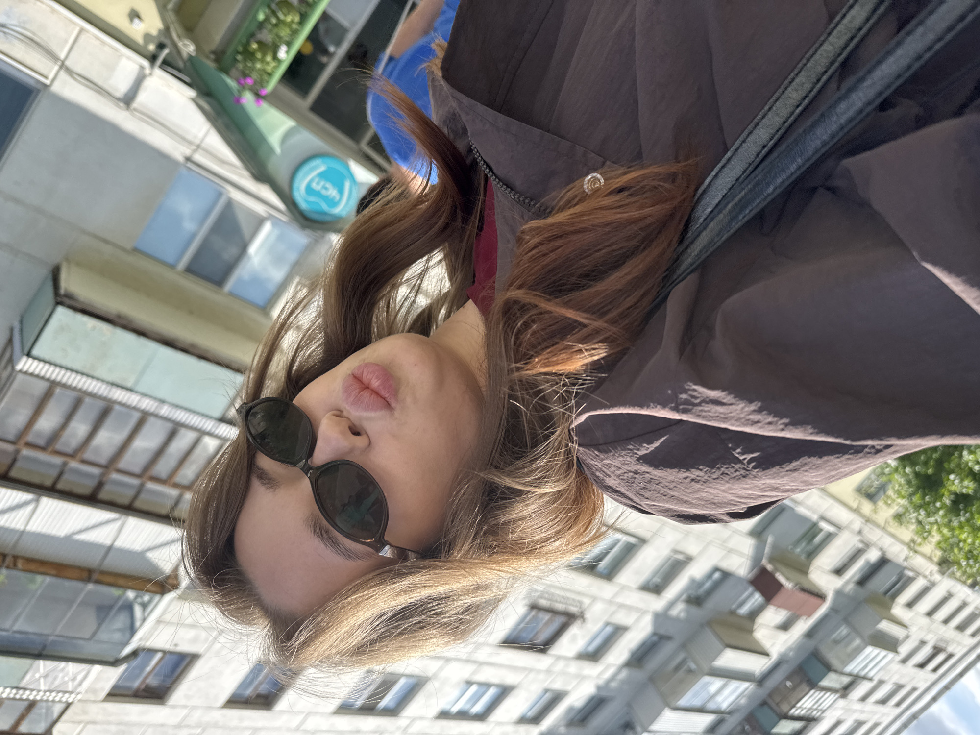 A selfie is taken from a slightly low angle by a young woman with long, light brown hair, wearing dark sunglasses and a dark brown jacket over a maroon shirt. She is puckering her lips. The background is an urban environment, featuring a light-colored building with many windows and a portion of a green awning with a blue circular logo. The building appears to be an apartment complex, consistent with the architectural style found in Chaikovsky, Russia. Sunlight is casting shadows, indicating it is daytime.