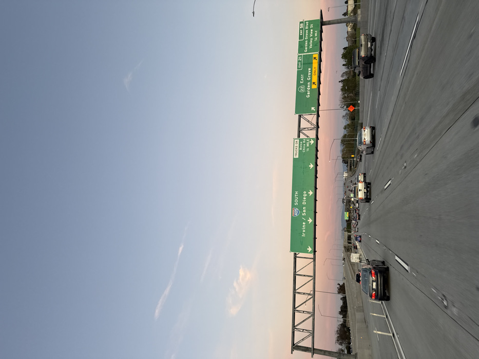 A highway stretches into the distance, filled with cars and trucks. Overhead, green signs indicate upcoming exits. One sign points south on the 405 towards Irvine and San Diego. Another sign indicates the 22 East towards Garden Grove, with an exit for Garden Grove Blvd and Valley View St. The sky transitions from blue to a soft pink hue near the horizon, likely indicating either sunrise or sunset.