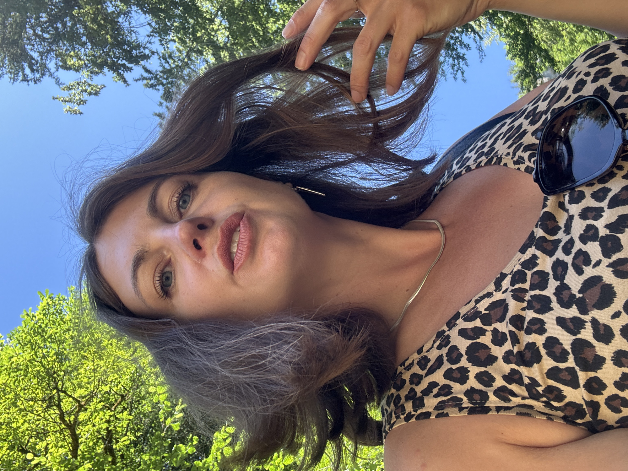 A woman, viewed from a low angle and from the shoulders up, is captured in an outdoor setting. She has brown hair, styled with some strands windswept, and is looking towards the camera with a slight smile. Her eyes are blue, and she is wearing makeup. A thin, gold-colored necklace is visible around her neck. She is dressed in a leopard-print tank top, and a pair of dark sunglasses is clipped to the front of her top. Her right hand is raised, gently touching her hair. Behind her, the bright blue sky is visible, interspersed with lush green trees and foliage. The lighting suggests it is a sunny day. The specific location in Saint-Claude, France, cannot be determined from the visual information provided in the image.