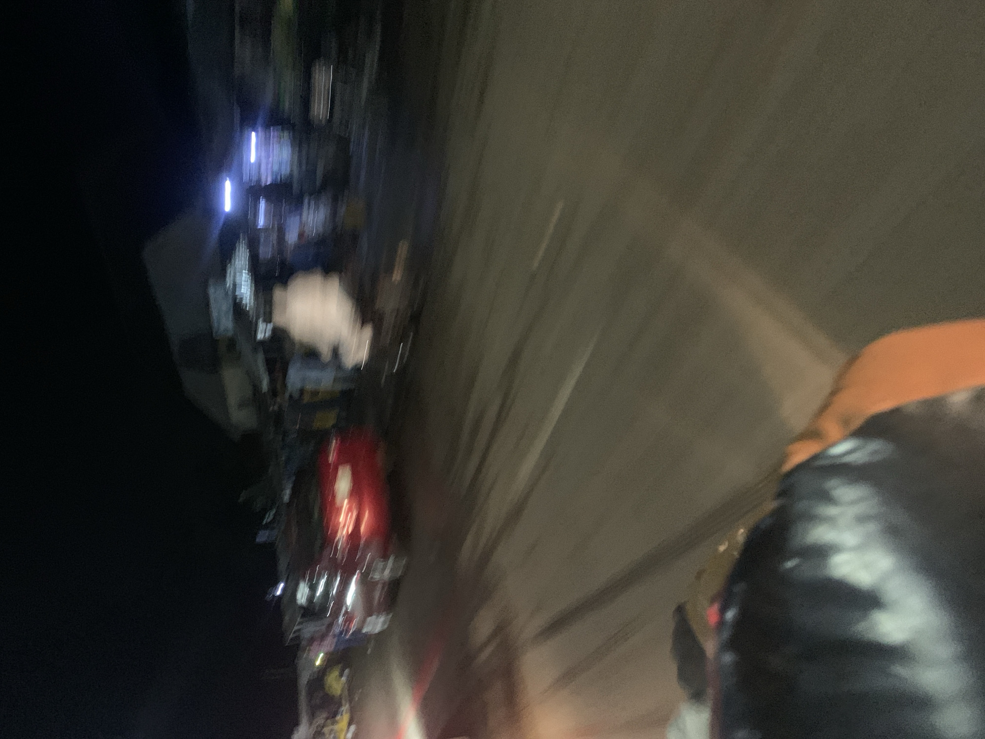 The image depicts a night scene in an urban environment, possibly on a street or road. The lighting suggests it's dark, with artificial lights from vehicles and buildings present. A red car is visible, its taillights illuminated, indicating it's either parked or in motion. Several indistinct figures, appearing as blurs due to motion, are present near the car and on what seems to be the side of the road, suggesting a public area or a gathering. The road surface shows tire marks, possibly from the movement of vehicles. The background includes structures that appear to be buildings, some with illuminated signs or lights. The foreground shows a blurred section of what could be the interior of a vehicle or an object positioned close to the camera, obscuring a portion of the scene. The overall impression is of a busy, possibly commercial or residential, area at night, with vehicular and pedestrian activity.