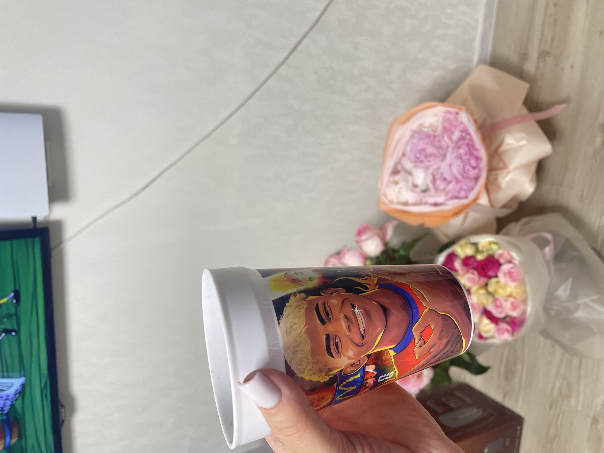 The image shows a hand with pale pink nail polish holding a white cup with a cartoon image of a smiling young man with blonde curly hair and braces. Behind the cup, there are two bouquets of flowers, one with pink and yellow roses in a white wrapper, and another with pink flowers wrapped in orange paper. In the background, a television screen is visible with a cartoon image, and a white wall with a wire running down it. The scene appears to be indoors, possibly a room in a residential building. There is no information provided to ascertain the location of Rozsoshentsi, Ukraine.