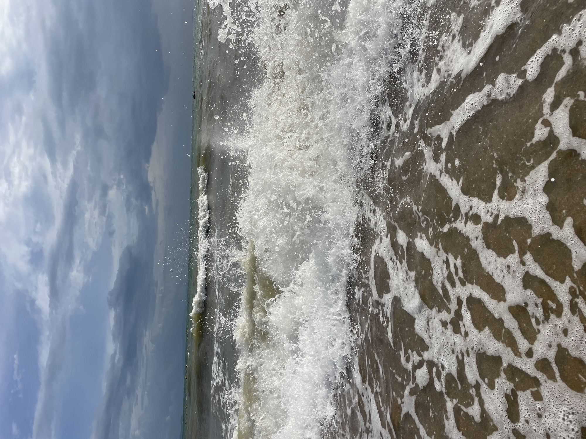 The image depicts a choppy sea with a breaking wave in the foreground, viewed from a low angle at the water's edge. The water is a murky green-brown, with white foam and spray from the wave. In the distance, there are other smaller waves and a dark, overcast sky. A small, dark object, possibly a boat, is visible on the horizon. The sand at the water's edge is wet and partially covered by the receding foam. The scene suggests a somewhat rough day at the beach, possibly during a storm or in choppy weather. The location is identified as Pattaya City, Thailand.
