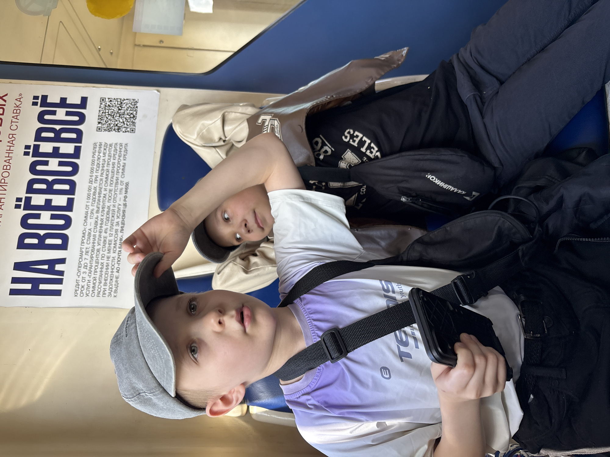 Two boys are seated in an interior space, likely public transport, in Kukmor, Russia. The boy in the foreground, estimated to be 6-10 years old, wears a grey baseball cap, a white and purple ombre athletic t-shirt with "TENNIS youth" and an "E" logo, and dark trousers with a black belt. He holds a black smartphone with a textured case in his right hand and has a black cross-body bag across his chest. His gaze is directed slightly to the right of the camera. The boy in the background, estimated to be 7-11 years old, wears a dark baseball cap, a black t-shirt with partial white text "ELLES" (possibly "LOS ANGELES"), and dark trousers. A light brown jacket with "86" and other symbols is draped over his shoulders. He also wears a black cross-body bag, prominently displaying "КАЛАШНИКОВ" (Kalashnikov) text. His attention is directed towards the first boy.
The interior features blue upholstered seats and light-colored walls. A white paper poster is affixed to the wall behind the boys. The poster has Russian text in blue, reading "ГАРАНТИРОВАННАЯ СТАВКА" (Guaranteed Rate) and large text "НА ВСЁВСЁВСЁ" (For Everything, Everything, Everything). Smaller text details a credit offer from "ПОЧТА БАНК" (Post Bank) with terms ranging from 1,000,001 to 6,000,000 rubles, 3 to 7 years, and rates from 17.9% annual, with a potential 4% annual rate under certain conditions, and mentions a license number (ЦБ РФ №650). A QR code is present on the poster. A reflection showing yellow and white objects is visible on a curved surface to the right.