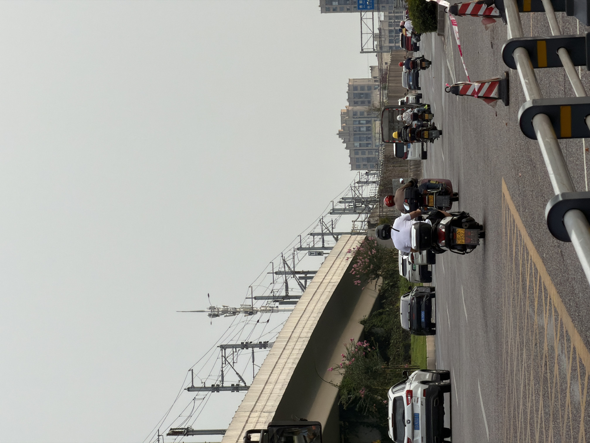 The image depicts a street scene in Jiangjin, China, with a focus on a multi-lane road under an elevated railway or highway structure. Several vehicles, including cars and motorcycles, are visible on the road. On the left, a white SUV with license plate "D N5074" is in the foreground. Further back, a black car with license plate "A J593" is also on the road. In the middle of the frame, several motorcycles are traveling, one with a rider wearing a white shirt and a black helmet, and another with a rider wearing a brown jacket and a red helmet. To the right, traffic cones with red and white stripes and caution tape are set up, indicating a potential road closure or construction. In the background, buildings and power lines are visible, characteristic of an urban environment. The sky is overcast, suggesting a cloudy day.
