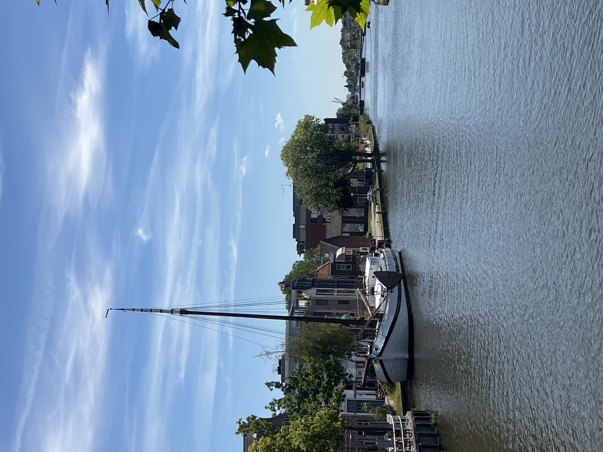 The image captures a serene canal scene in Delft, Netherlands, on a bright, clear day with a blue sky dotted with wispy white clouds. A single, tall-masted sailboat, predominantly white with a dark hull, is moored along the left bank, its mast reaching high into the sky. The boat appears to be unoccupied. The left bank of the canal is lined with a row of historic buildings, their facades displaying a mix of traditional Dutch architecture with varying rooflines and window styles. Lush green trees are interspersed between the buildings, providing shade and adding to the picturesque setting. A black metal dock with a railing is visible to the left of the sailboat.
The canal itself is wide and calm, reflecting the sky and the surrounding environment. In the distance, the canal continues, lined with more buildings and trees, and a bridge spans across it. Further in the background, cranes and other structures suggest the presence of a more industrial or port area. The overall atmosphere is peaceful and idyllic, typical of a Dutch canal town. There are no people clearly visible in the immediate vicinity of the boat or the buildings on the left bank. However, in the distance, on the right bank, there are a few figures walking. The water's surface is slightly rippled, indicating a gentle breeze. Green leaves from a nearby tree frame the top right corner of the image, adding a natural element to the composition.