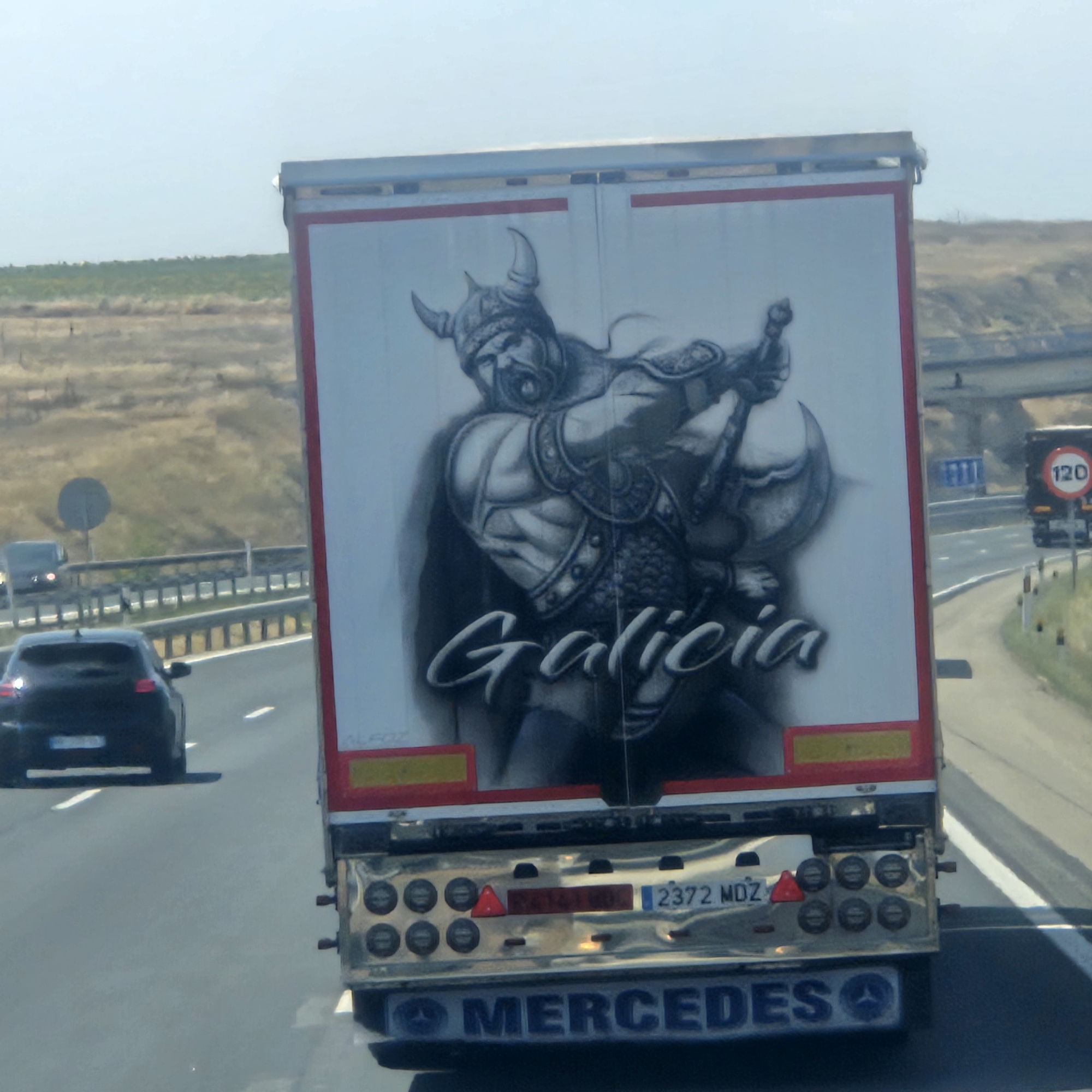 The image shows the rear of a white semi-trailer truck on a highway. The trailer is decorated with an airbrushed mural of a Viking warrior with a helmet and beard, holding a shield and axe. The word "Galicia" is painted in large, stylized script below the warrior. The truck has a chrome rear bumper with several round lights and a license plate that reads "2372 MDZ". The Mercedes-Benz logo is visible on the lower part of the rear of the truck.

The truck is in motion on a highway, with other vehicles visible in the background, including a black car in the left lane and another truck with a "120" speed limit sign in the right lane. The environment appears to be rural or semi-rural, with dry, rolling hills and vegetation visible on either side of the highway. The sky is clear and bright, suggesting a sunny day. The image appears to have been taken from another vehicle also traveling on the highway. The location is identified as Santa María Ribarredonda, Spain, which is a municipality in the province of Burgos, Castilla y León.