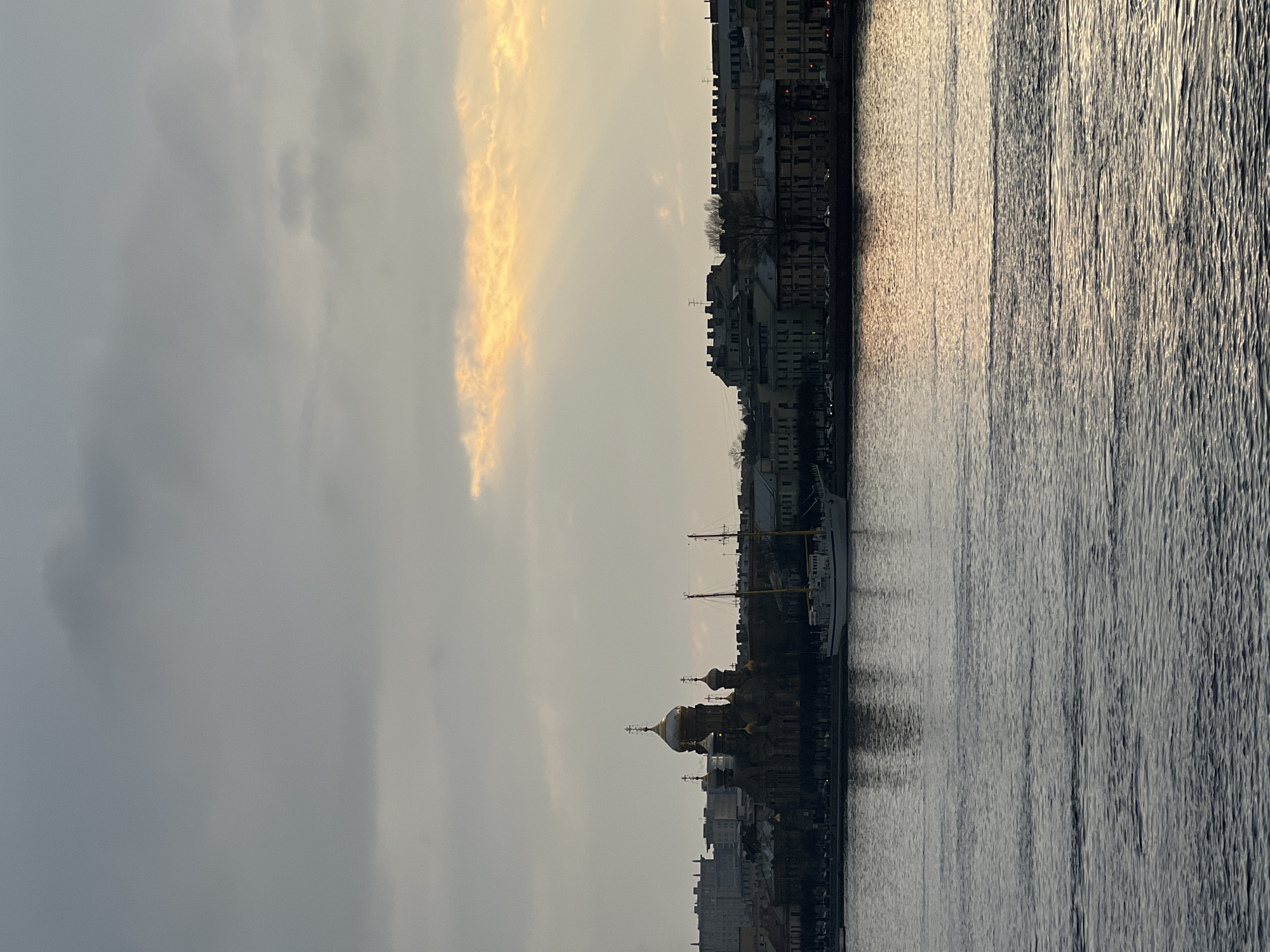 The image presents a cityscape along a body of water. On the right, the water reflects sunlight, creating a shimmering effect. Adjacent to the water is a row of buildings, densely packed together, indicating an urban environment. Some buildings are topped with golden domes, suggesting the presence of a church or cathedral. In the background, the sky is overcast with clouds, some tinged with a golden hue, possibly from the setting or rising sun.
Based on the architecture, particularly the golden domes and the row of historic buildings, the location is likely Saint Petersburg, Russia. The water body is likely the Neva River, a major waterway flowing through the city. The domed structure is likely the Smolny Cathedral.