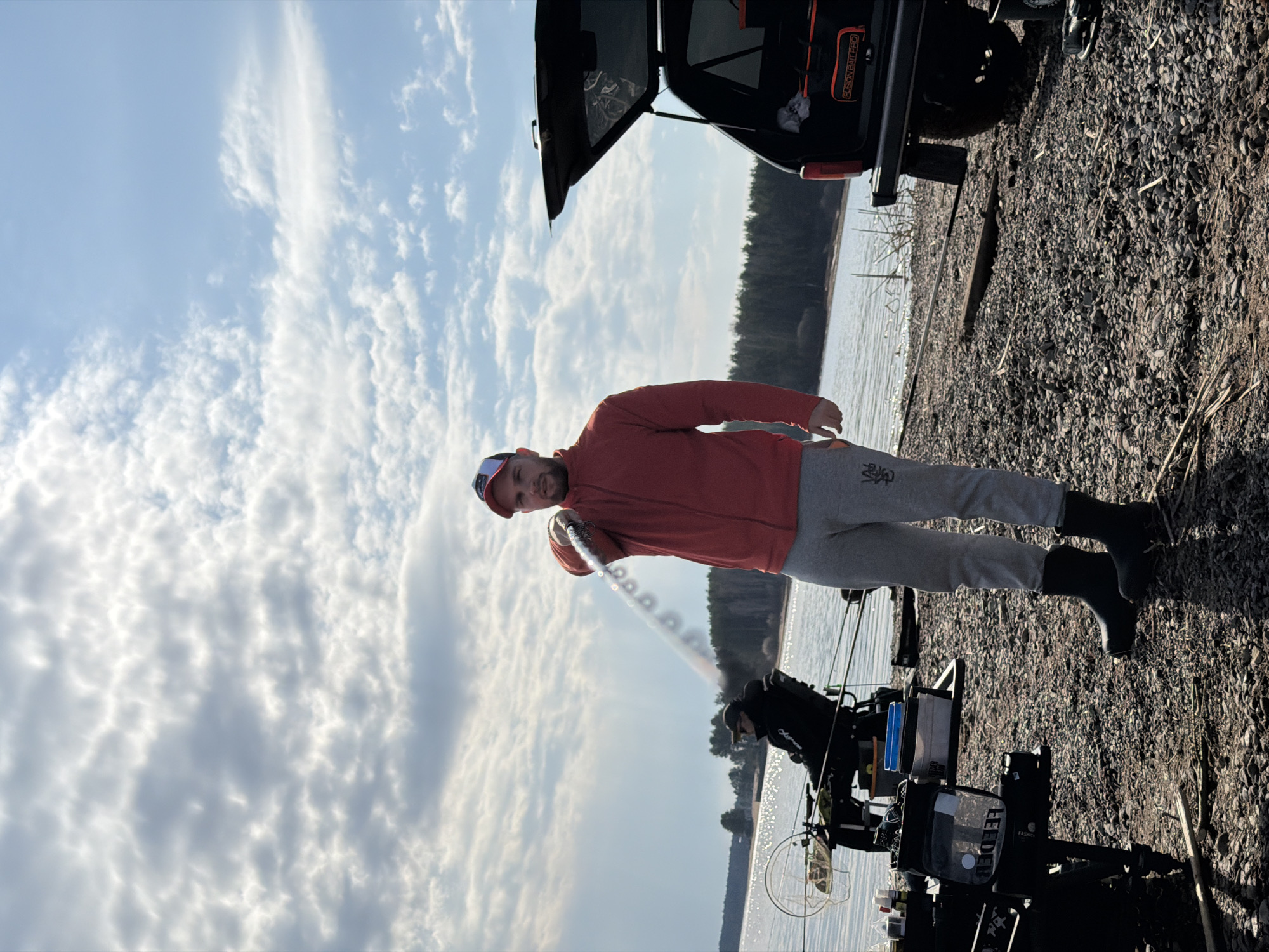 An outdoor scene in Chaykovsky, Russia, captures a man standing by a large body of water, likely engaged in fishing. The man, wearing a red long-sleeved shirt, grey sweatpants, and a baseball cap, is holding a long, white telescopic fishing rod over his shoulder. He stands on a stony, gravelly shore next to an array of fishing equipment.
To the left, a dark-colored vehicle with its trunk open is parked on the shore. Inside the trunk, an orange bag with the text "FUSION BAIT PRO" is visible. Near the water's edge, various pieces of fishing gear are set up, including multiple rod holders, a seat or platform, and a transparent container labeled "LEEDER" (possibly "FEEDER" with a stylized font). Another person or equipment is partially visible further down by the water.
The background features a calm body of water extending to a distant tree line. The sky is bright and partly cloudy, with a mix of fluffy white cumulus and thin wispy clouds, indicating daylight, likely late morning or early afternoon. The ground is covered in small stones and pebbles, with some dry reeds or grass scattered about. The man appears focused and calm in the outdoor setting.