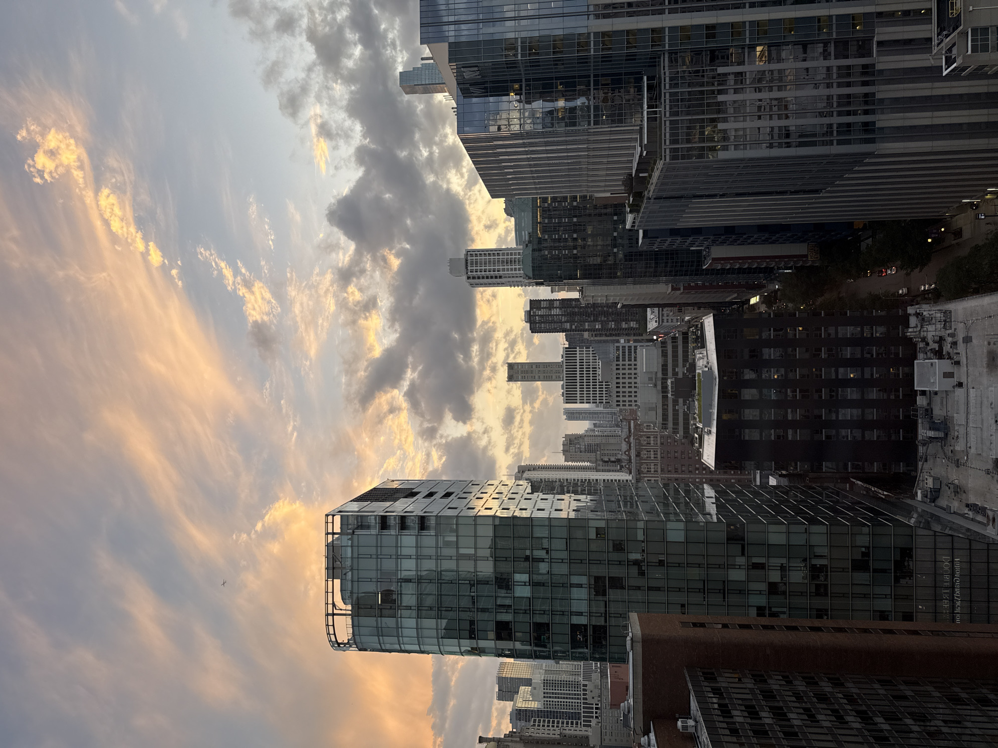 This is an aerial photograph of a cityscape at dusk, likely in Chicago, United States of America. The sky is filled with clouds, some illuminated with hues of orange and pink from the setting sun, while others are darker and more ominous. A small airplane is visible in the upper left quadrant of the sky. The foreground is dominated by the reflective glass facades of modern skyscrapers, which are interspersed with older brick buildings and more utilitarian structures. Some windows in the buildings are illuminated, suggesting that people are inside. The street level is not clearly visible, but hints of cars and streetlights indicate urban activity. The overall impression is one of a bustling metropolis transitioning from day to night under a dramatic sky.