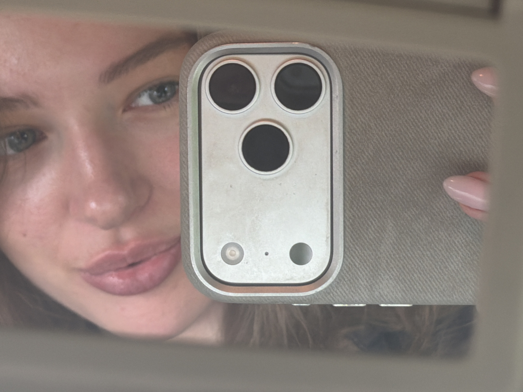 A close-up, mirrored selfie shows a young woman's face, specifically her eyes, nose, and lips, reflected in a phone camera lens. The phone is held by her hand, with two pink manicured fingernails visible on the right side of the frame. The phone has a textured gray case and a distinctive triple-lens camera array on the back, with three circular lenses arranged in a triangular formation. The background appears to be a textured surface, possibly the interior of a car. The lighting suggests an indoor setting. The woman's facial features are soft and lightly made up. The overall image is candid and informal.