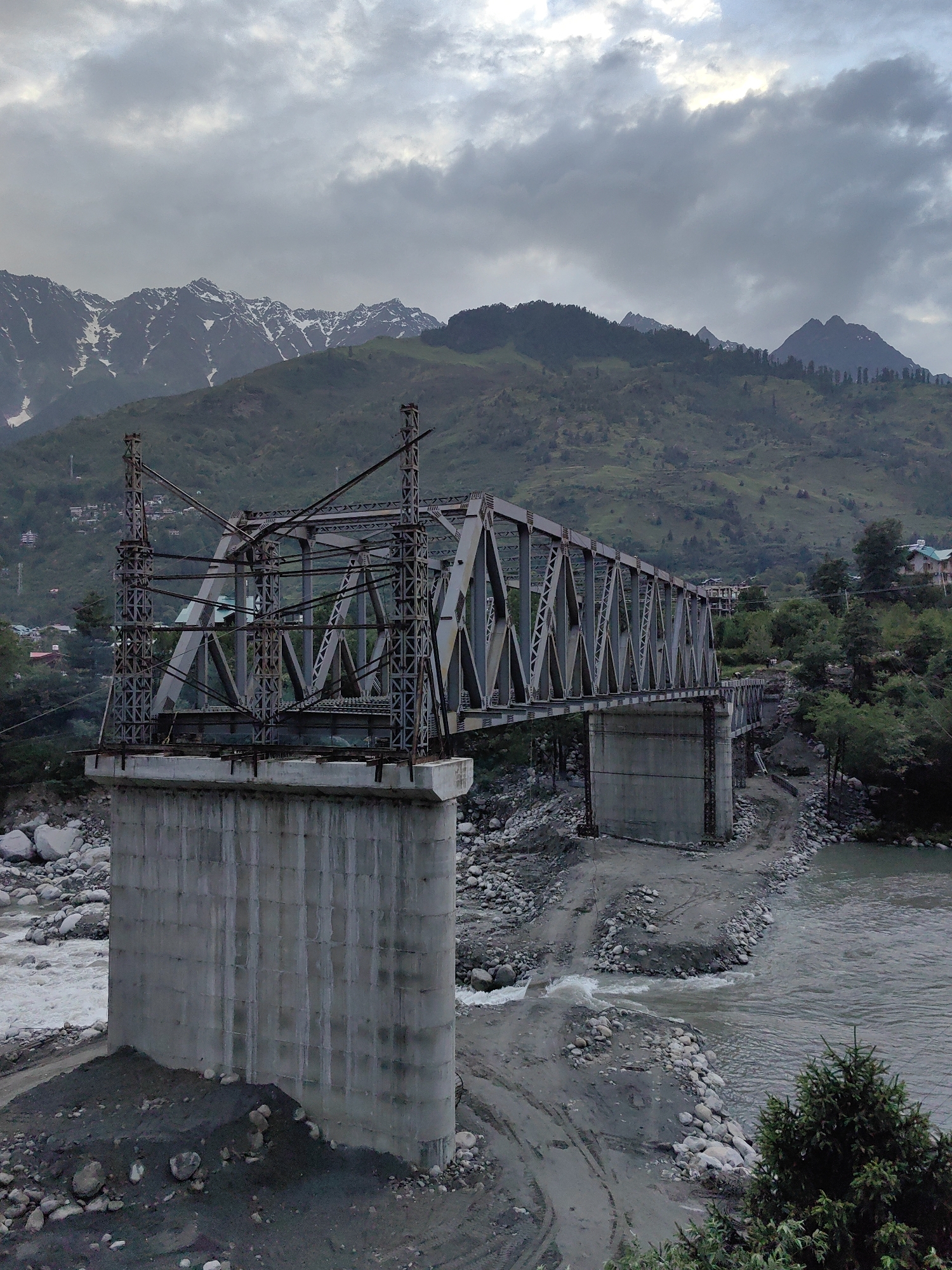 The image depicts a large metal truss bridge under construction, spanning a rocky riverbed with a shallow, flowing river. The bridge is supported by massive concrete pillars. The scene is set in a mountainous region, with lush green hills and snow-capped peaks visible in the background. The sky is overcast with dramatic, dark clouds, suggesting a cloudy or stormy day. The overall mood is one of rugged natural beauty and ongoing development. The location appears to be Kulang, India, as indicated by the context. There is no visible text in the image.
