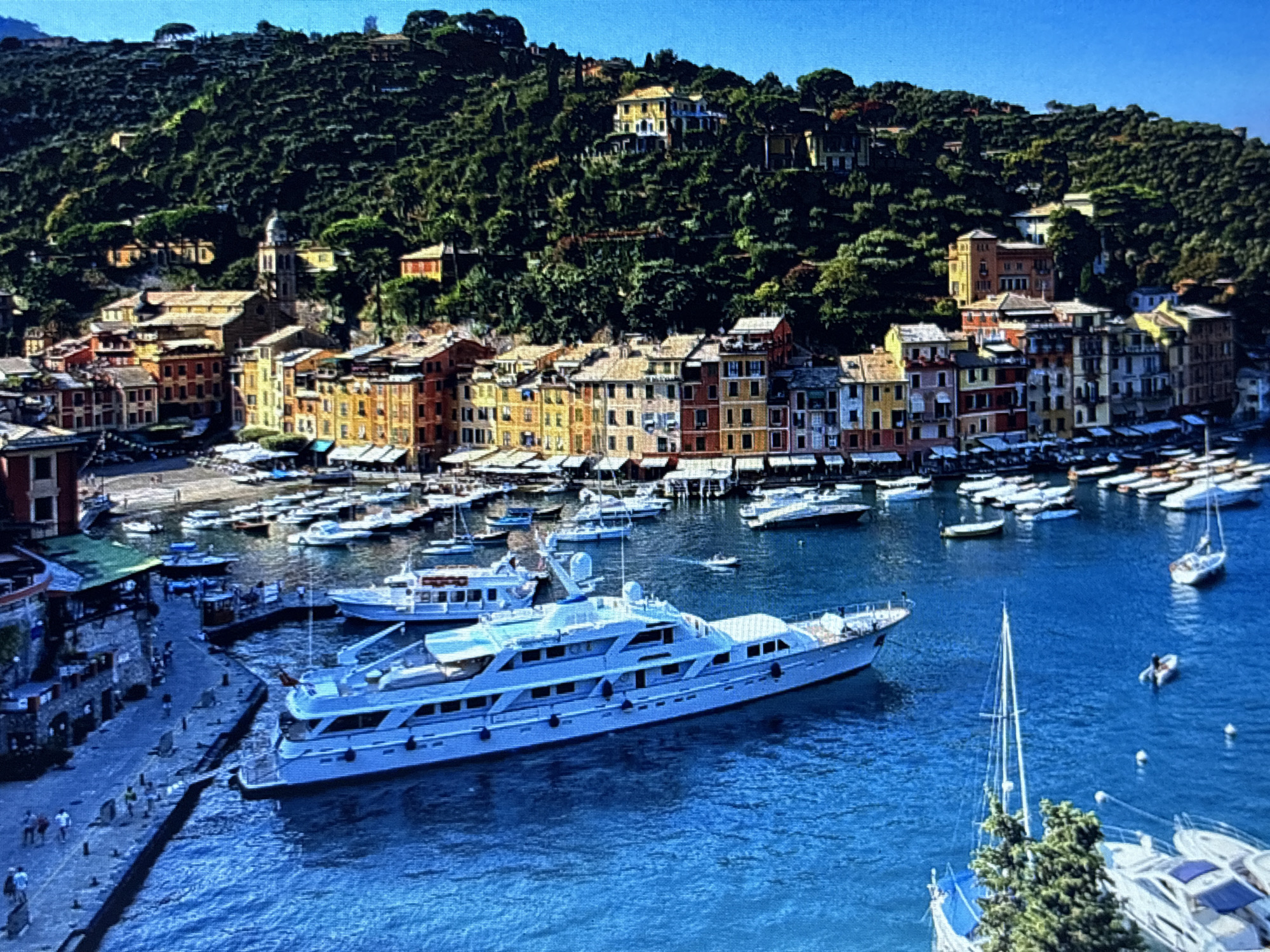 This media file captures a picturesque harbor scene in Portofino, Italy, under a clear blue sky, suggesting a sunny day. The image is dominated by a large, elegant white yacht prominently positioned in the foreground, its sleek lines and multiple decks indicative of luxury. Surrounding the yacht are numerous other boats, including smaller yachts and sailboats, moored in the vibrant blue water.
The background reveals a charming Italian coastal town characterized by colorful buildings with terracotta roofs lining the waterfront. These structures are nestled against a lush, green hillside densely covered with trees. Several buildings are visible higher up the hill, some appearing to be large villas or homes, suggesting a wealthy and scenic setting. A church with a distinct dome and bell tower stands out amongst the town's architecture.
Along the stone quay in the left foreground, several people are visible strolling and gathered, adding a sense of life and activity to the scene. The overall atmosphere is one of tranquility and leisure, typical of a Mediterranean resort town. No visible text is present in the image. The time of day appears to be midday, given the bright sunlight casting shadows.
