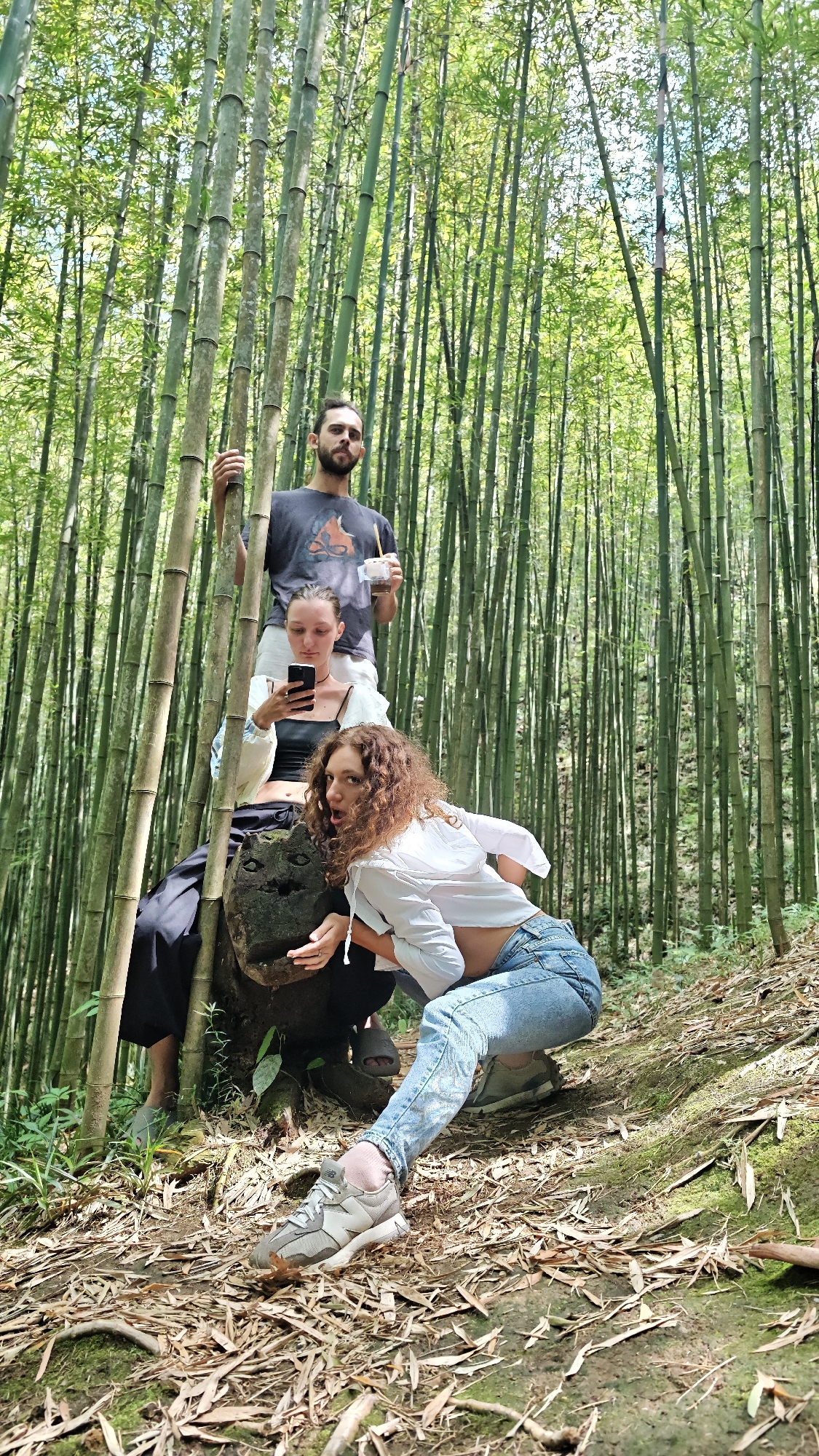 In a bamboo forest located near Ngã Ba Kim, Vietnam, a man stands holding bamboo stalks while holding a drink. A woman sits below him, looking at her phone. Another woman in a white top and jeans squats in front of them, looking at the camera with a playful expression. The women are sitting on or near a carved, weathered stone. The forest is dense with tall, green bamboo stalks. The ground is covered with dry leaves and moss.