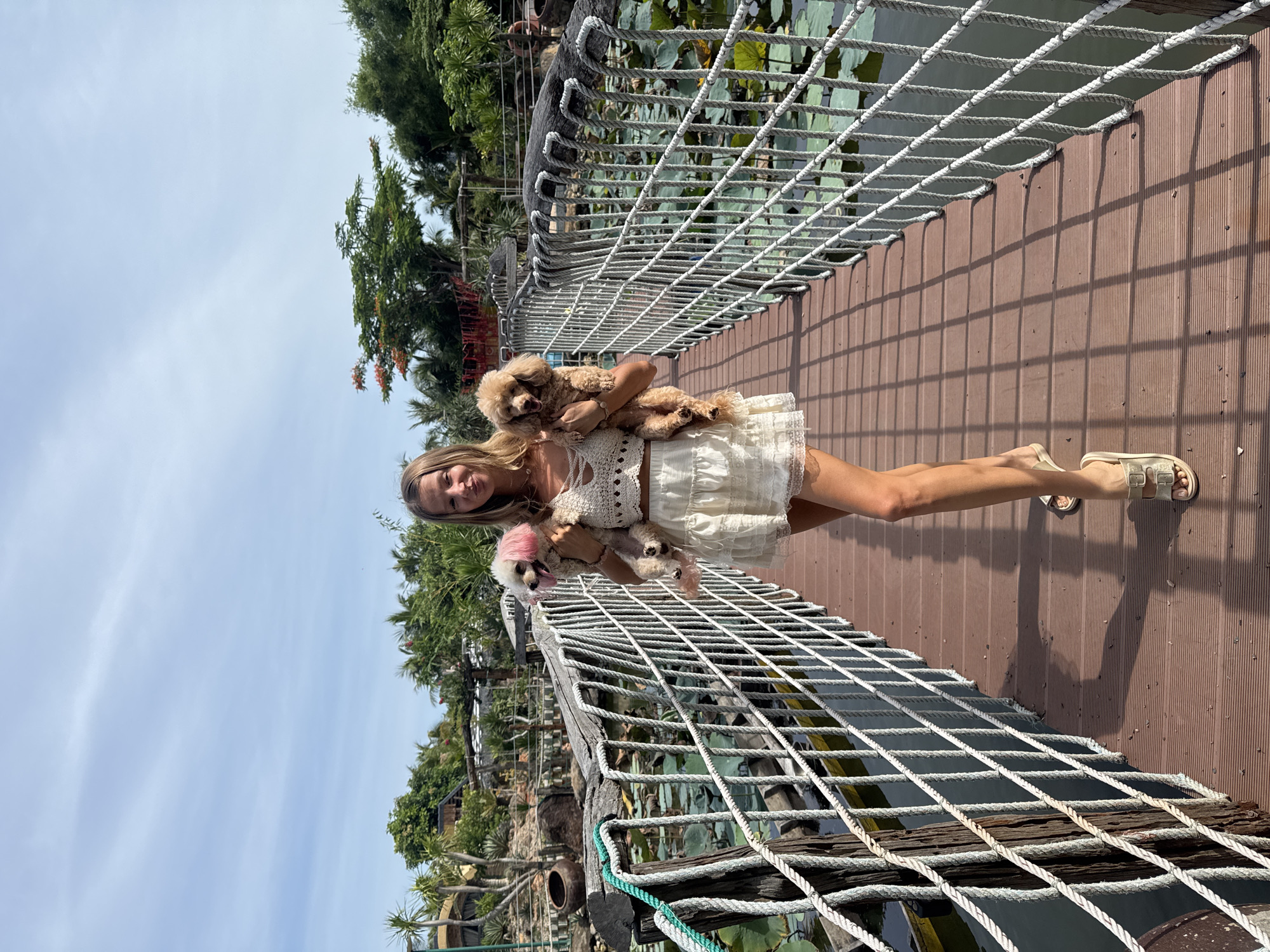 A young woman stands on a wooden bridge over a pond covered with lily pads. She is wearing a white crochet top and a white ruffled skirt. She is holding three small dogs: one in her left arm with pink-dyed fur on its ears, and two in her right arm. The bridge has rope railings. The background shows lush green trees and vegetation, suggesting a tropical environment. The sky is partly cloudy. The scene appears to be in Mũi Né, Vietnam, a coastal resort town known for its beaches and sand dunes.