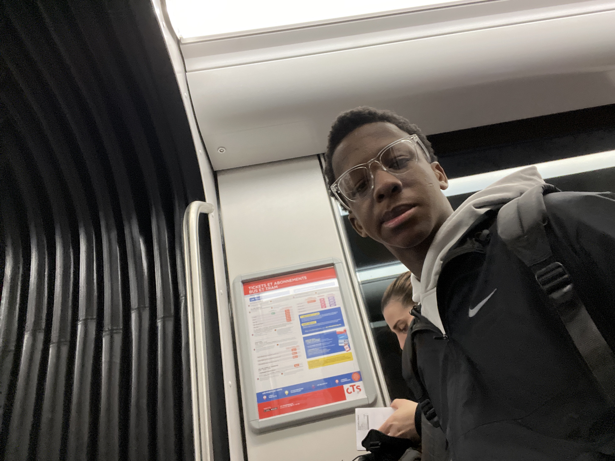 The image shows a young man wearing glasses and a black Nike jacket standing inside a Strasbourg CTS public transportation vehicle, likely a tram or bus. A woman is standing behind him, partially visible. An advertisement for CTS bus and tram tickets is mounted on the wall. The interior features corrugated black paneling.