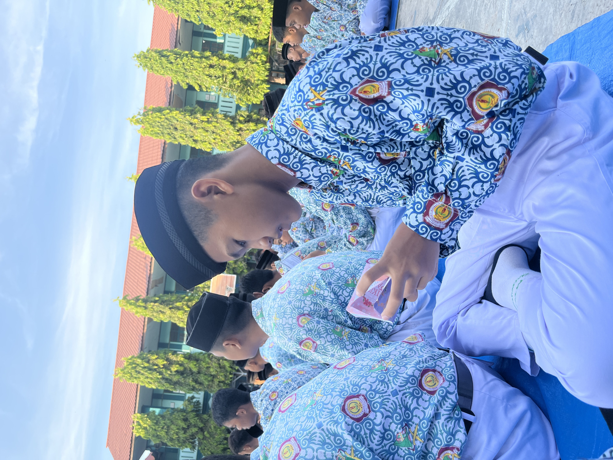A group of young male students wearing white pants and patterned blue shirts are seated outdoors. The students are wearing black peci hats. One student in the foreground is holding a small, pink object, possibly a booklet or card, and looking down at it. The students appear to be in a school setting, with a building and trees visible in the background. The sky is clear and blue. The image was taken on October 20, 2023. The students' attire suggests a religious or cultural event. The pattern on their shirts includes a circular emblem with text. The location is the courtyard of a school, possibly a Madrasah Tsanawiyah or a similar Islamic educational institution in Lhokseumawe, Indonesia.