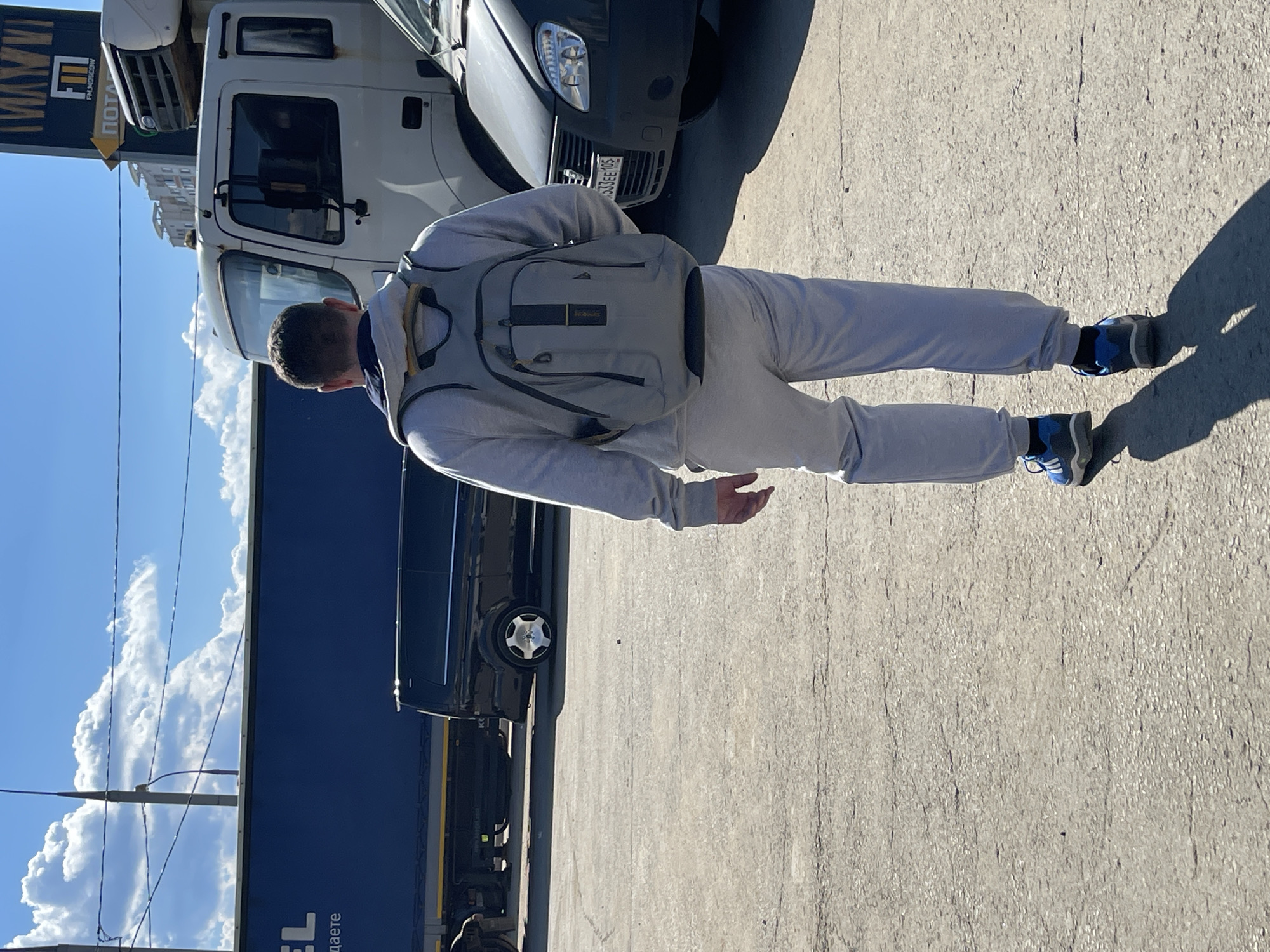 This full-length outdoor image captures a person from behind, standing on a light gray concrete or asphalt surface under a clear blue sky dotted with fluffy white clouds. The person is wearing a light gray hooded sweatshirt, matching sweatpants, and a gray backpack with yellow accents. They have dark, short hair and are wearing blue and gray sneakers. Bright sunlight casts strong, distinct shadows, indicating a sunny daytime scene.
In the background, a commercial or industrial setting is visible, consistent with Moscow, Russia. Directly behind the person is the white cab of a large truck. To the right of the person's lower body, the front bumper and headlight of a dark gray passenger car are partially visible, bearing a license plate that reads "333EE 10S". To the left of the person, a large dark blue truck trailer displays partial white Cyrillic text, including "EL" and "даете". Above the white truck, a yellow and black sign with a downward-pointing arrow clearly shows the Cyrillic letters "MKM" and "ПОСТАВЩИК," which translates to "Supplier."