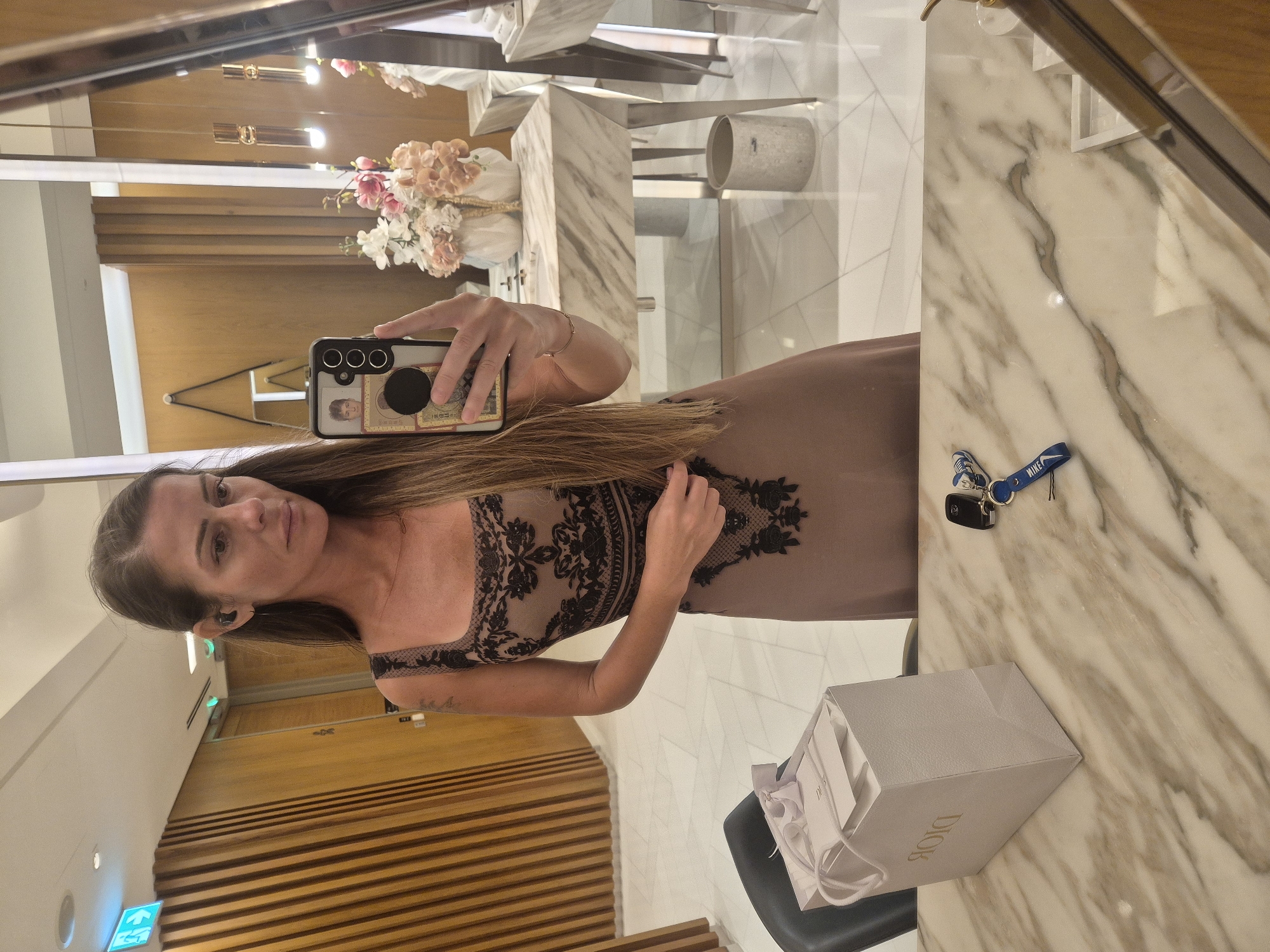 A woman is captured in a mirror selfie within an indoor setting, likely a well-appointed restroom or dressing room. She has long, dark brown hair and is wearing a form-fitting dress in a taupe or light brown color, featuring intricate black lace-like patterns on the bodice and upper skirt. An earbud is visible in her left ear, and she has a simple gold bracelet on her right wrist. She holds a smartphone, possibly a Samsung model, with a clear case that reveals a pop socket, a picture or card, and what appears to be currency on the back. Her expression is neutral as she looks towards the camera.
The background behind her reflects a modern interior. To her right, a light-colored marble counter is visible, on which a set of car keys with a blue "NIKE" branded lanyard rests. Next to the counter, a light grey "DIOR" shopping bag sits on a black chair. Further in the background, a vase filled with artificial pink and white flowers is placed on the marble surface, and part of a metallic trash can is reflected. The room features horizontal wooden slatted walls, white tiled flooring, and modern rectangular light fixtures. A green illuminated exit sign is also visible on a plain wall to the left. The scene suggests a refined or upscale environment.