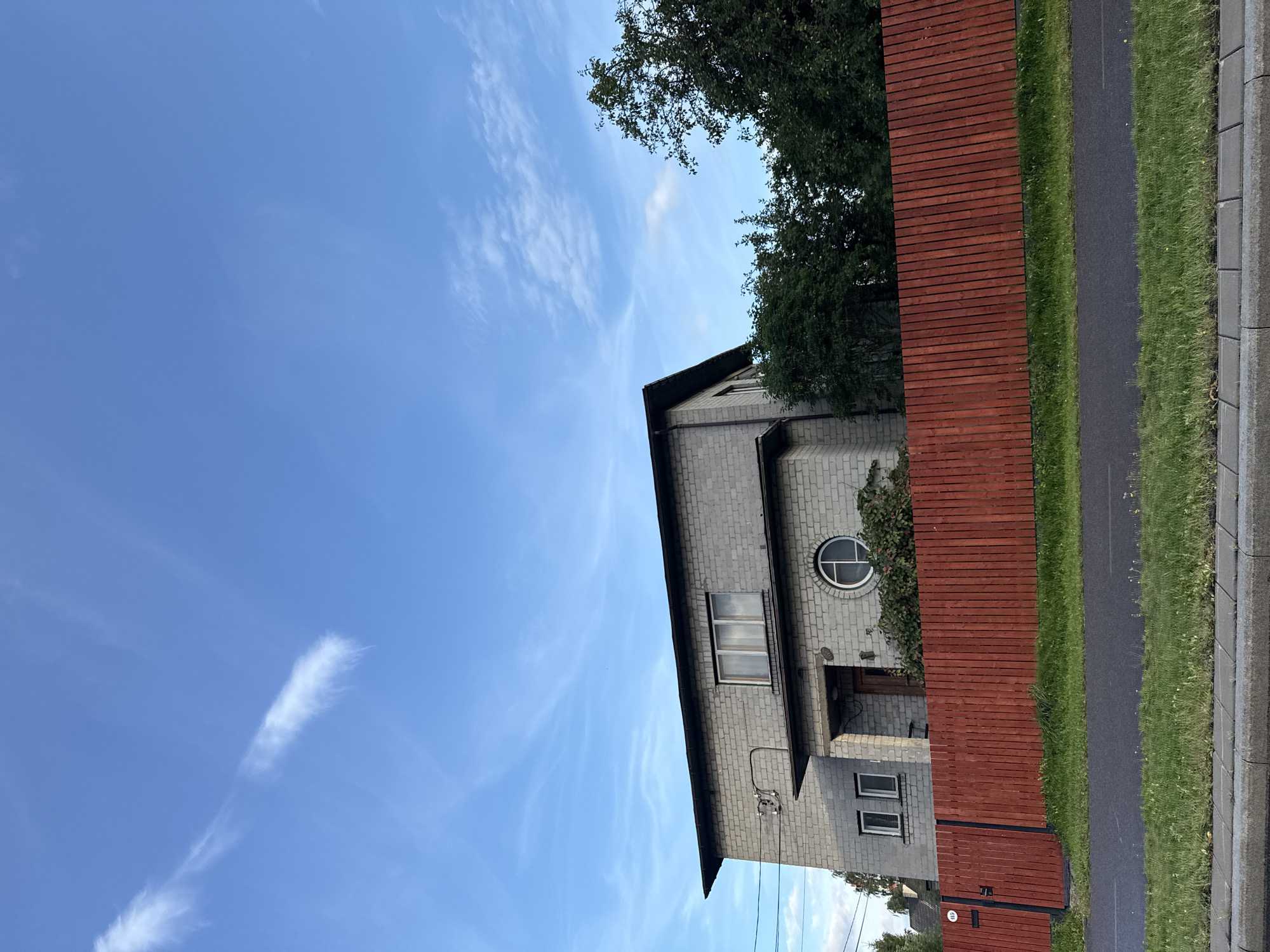 A two-story, light grey brick house with a dark sloped roof is visible under a bright blue sky with wispy white clouds. The house has a modern design with simple rectangular windows, and a distinctive circular window with a white frame on the first floor. A red wooden fence runs horizontally across the middle of the image, separating the property from a paved path and a strip of grass. The fence has vertical planks and a black metal gate on the left side, with a house number '145' visible on it. Trees and greenery are visible behind the fence, on the property. The overall scene suggests a residential area in a city like Tallinn, Estonia, likely during daylight hours in warmer weather. There are no people or animals visible in the image.