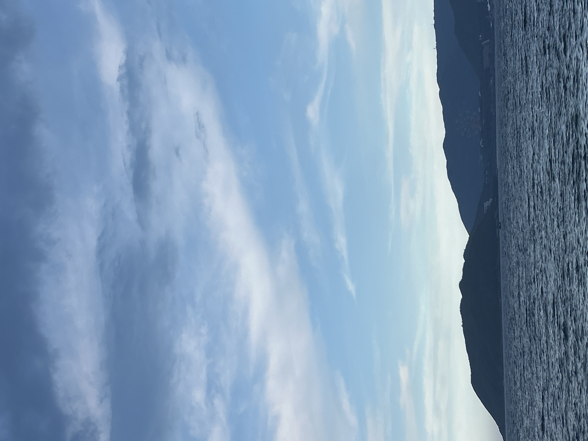 The image depicts a serene seascape with a prominent mountain range in the background. The sky is a mix of blue and grey clouds, suggesting a partly cloudy day. The sea is a deep blue, with gentle ripples indicating calm conditions. The silhouette of a hilly island occupies the left side of the frame, while the mainland with more rugged mountains stretches across the horizon on the right. There are no people or active subjects visible in the scene. The environment appears natural and undeveloped, with the mountainous terrain meeting the coastline. The scene is likely set in a coastal area of Nha Trang Ward, Vietnam, characterized by its beautiful natural landscapes.