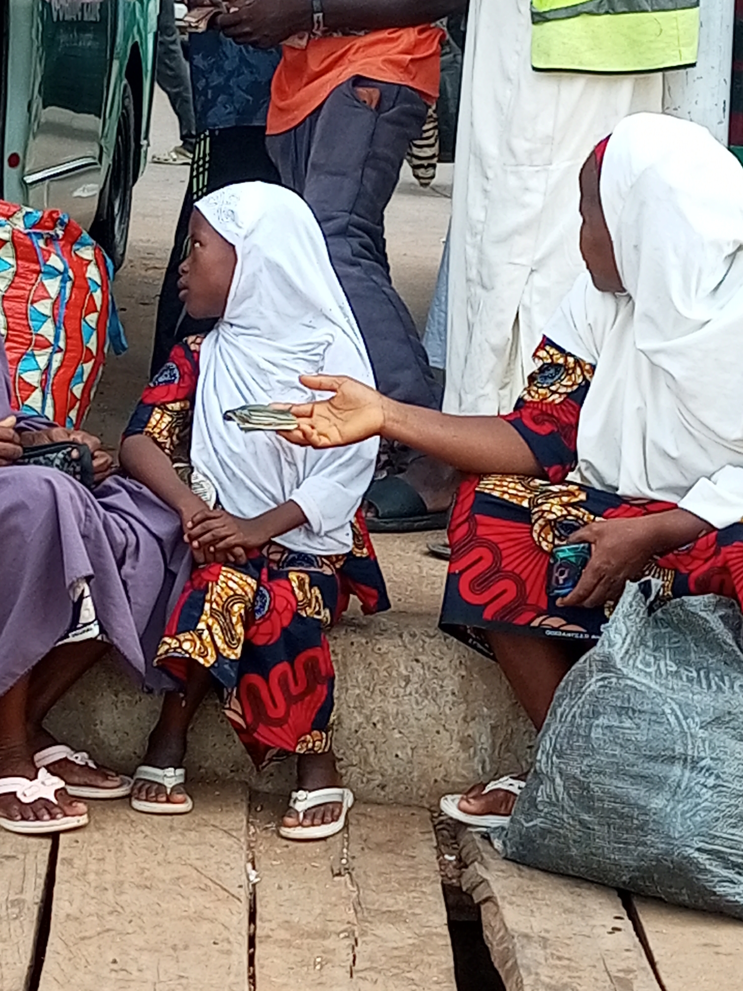 In Gombe, Nigeria, a child wearing a white hijab and a vibrantly patterned dress sits on concrete steps. The child holds a stack of banknotes. To the child's right, an adult, also in a white hijab and a matching dress, extends their hand, and their fingers are touching the banknotes. The adult is holding a small, green object in their other hand, and they are partially obscured by a large, grey, plastic bag. To the child's left, another individual in a purple garment is visible, seated with their legs crossed. In the background, a green vehicle and other people are present, suggesting a public area or market.