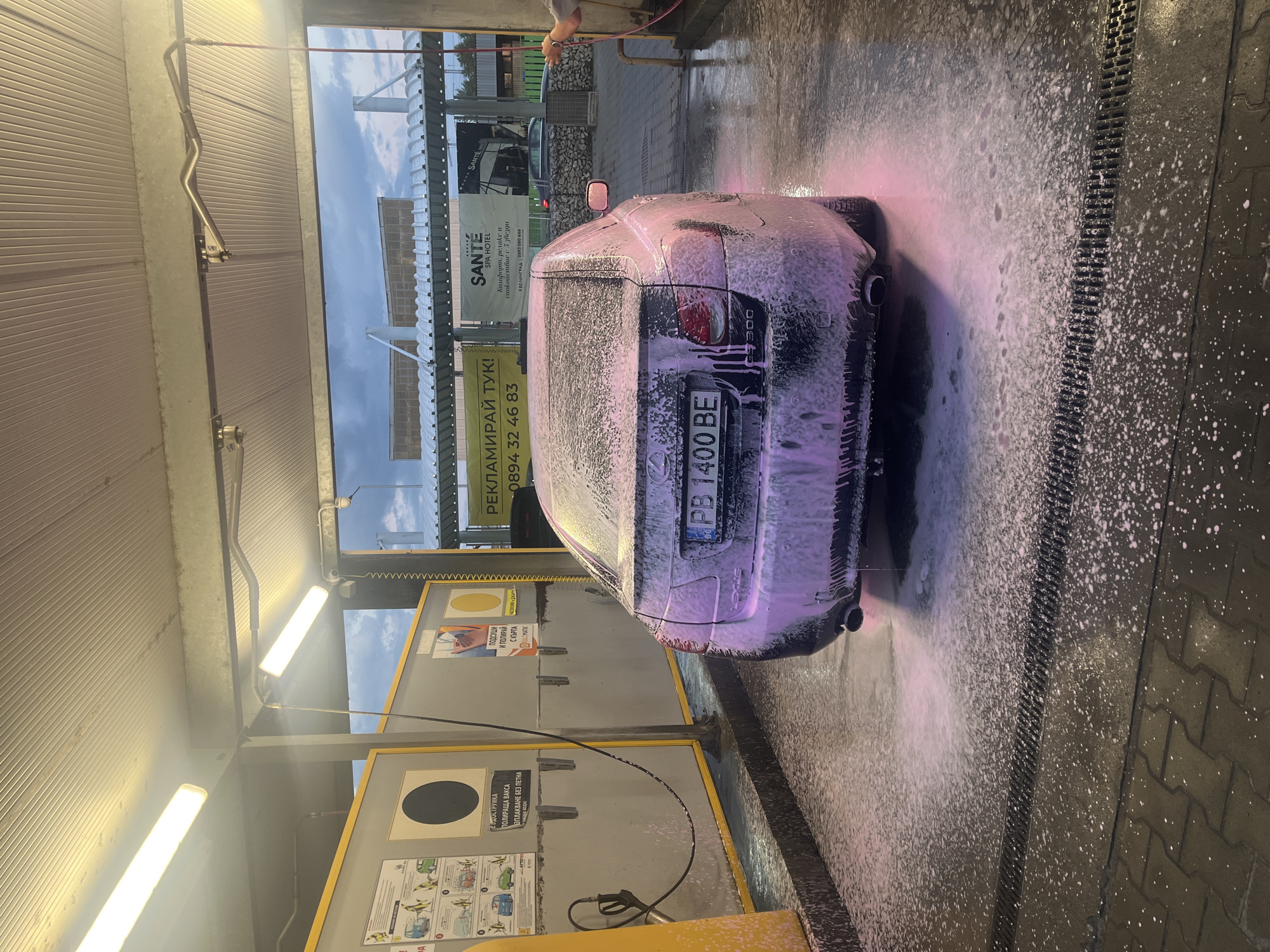 The image shows the rear of a dark-colored Lexus IS 300 car being washed at a self-service car wash station. The car is covered in pink soap foam. The license plate reads "PB 1400 BE." To the left of the car, there is a pay station with instructions in Bulgarian. Above the car, there is an advertisement that reads "РЕКЛАМИРАЙ ТУК! 0894 32 46 83". Another sign mentions "SANTÉ SPA HOTEL" in Velingrad. A person's hand holding a hose can be seen at the top right. The surrounding environment consists of a covered car wash bay with artificial lighting and drainage grates on the floor.