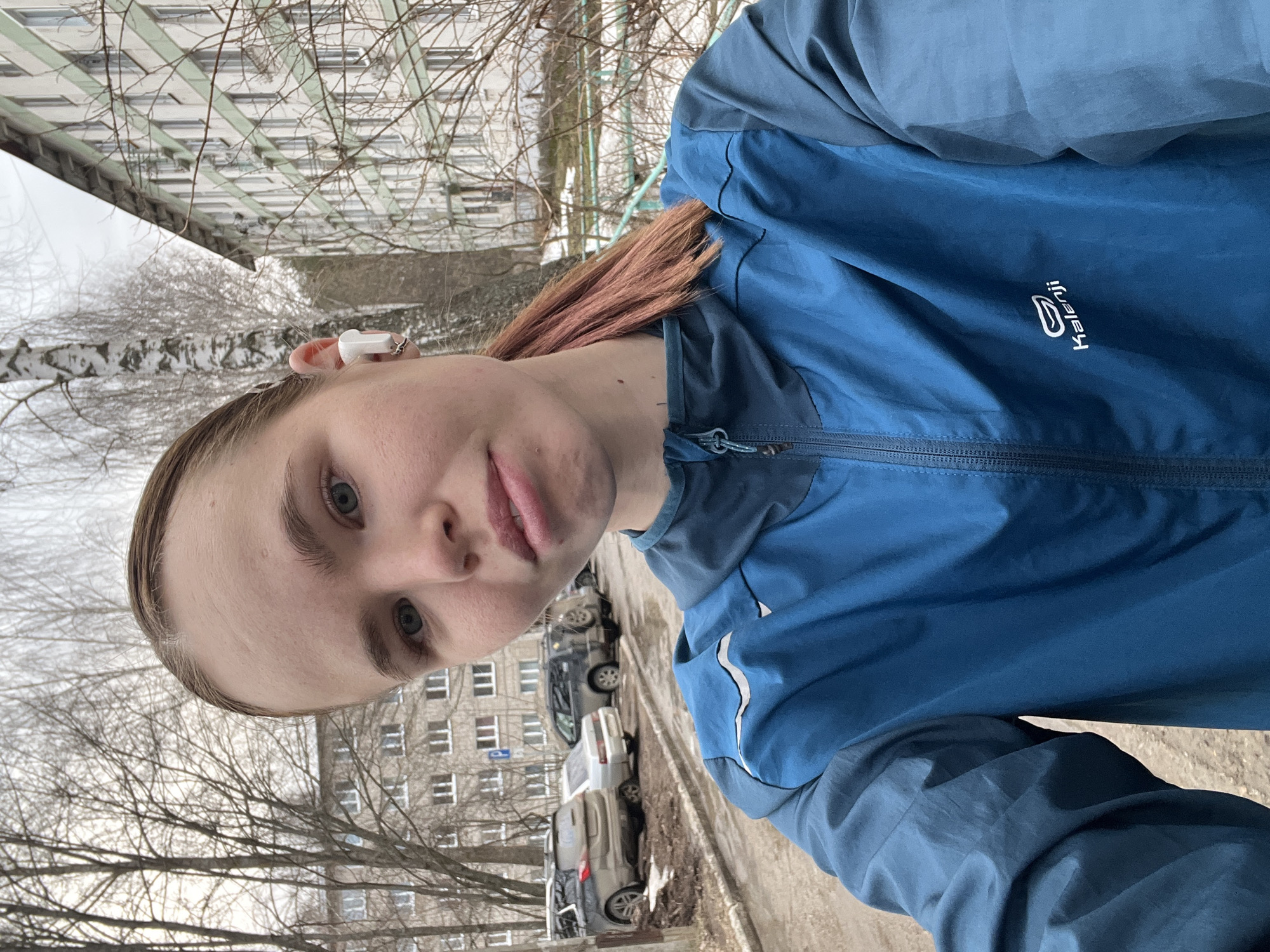 A selfie of a young person in a blue Kalenji jacket is taken outdoors, with a gray apartment building typical of Soviet-era construction visible in the background. Trees are bare of leaves, suggesting late autumn or early spring. Several cars are parked along the side of the road next to a concrete curb and some snow. The person is wearing a white earbud. The photograph appears to be taken in the city of Chaikovsky, Russia, as indicated by the architecture and overall urban environment.