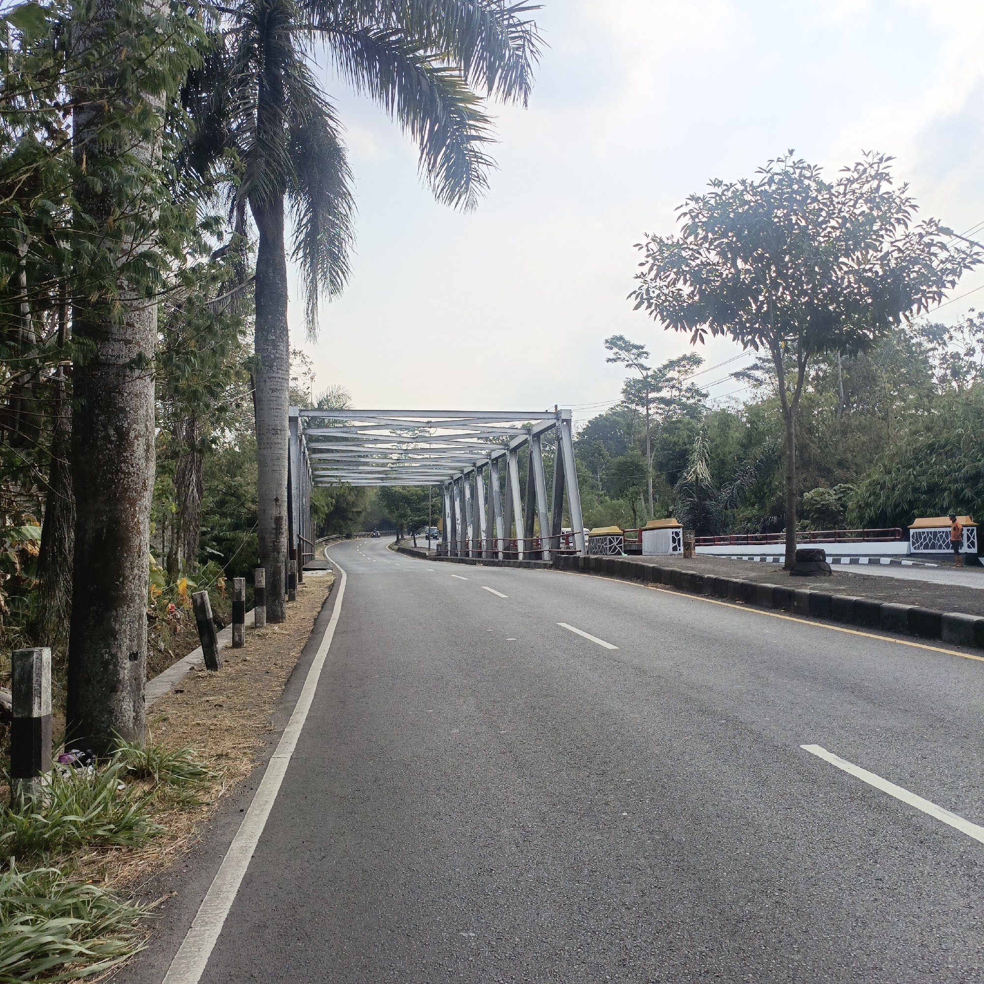 This image captures a view of a road leading towards a steel truss bridge, likely located in Salatiga, Indonesia, given the typical infrastructure and vegetation. The road is asphalt, marked with white lines indicating lanes. On the left side of the road, there are several large palm trees and other dense foliage, with concrete posts lining the edge of the road. The steel bridge has a distinct triangular lattice structure supporting its deck. Beyond the bridge, the landscape is characterized by more trees and vegetation, with a light-colored sky suggesting daytime. A few vehicles, including what appears to be a car and motorcycles, are visible on the road approaching the bridge. On the right side of the road, near the bridge, there are decorative barriers with black and white patterned elements. The overall scene depicts a rural or semi-rural setting with a functional piece of infrastructure.