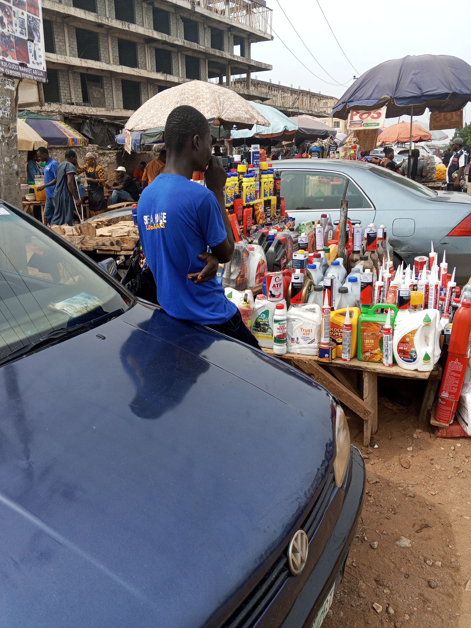 The image depicts a street market scene in Gudu Market, Abuja, Nigeria. A man in a blue shirt sits on the hood of a dark blue Volkswagen, positioned adjacent to a makeshift wooden table overflowing with automotive fluids and lubricants. The table is laden with various bottles, cans, and tubes of products, including brands such as "Trust ATF," "Golden Super," and "Rondo." Behind the table, a silver sedan is visible amongst the market stalls. The background reveals a partially constructed concrete building. Market activity is visible under colorful umbrellas. A POS sign indicates cash withdrawal and bill payment services. Debris litters the ground. A sign displaying "K35 GUDU MARKET ABUJA" is partially visible on a pole.