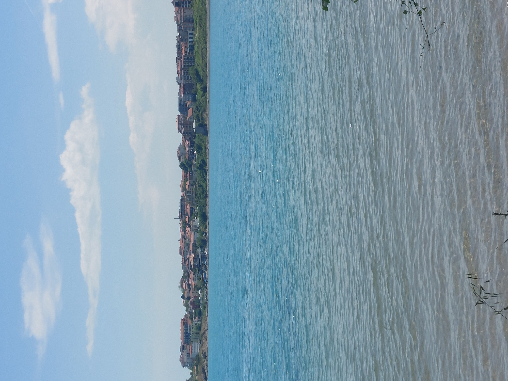 The image shows a shoreline in Tsarevo, Bulgaria, with the sea taking up the majority of the frame. The water transitions from a deeper blue further out to a lighter, more reflective tone near the shore, with visible ripples. A line of buildings and greenery stretches along the coastline on the left side of the frame. These buildings appear to be a mix of residential and potentially commercial structures, with predominantly red-tiled roofs. On the left is a blue sky with some scattered clouds. There are some bushes in the bottom corner on the right side.