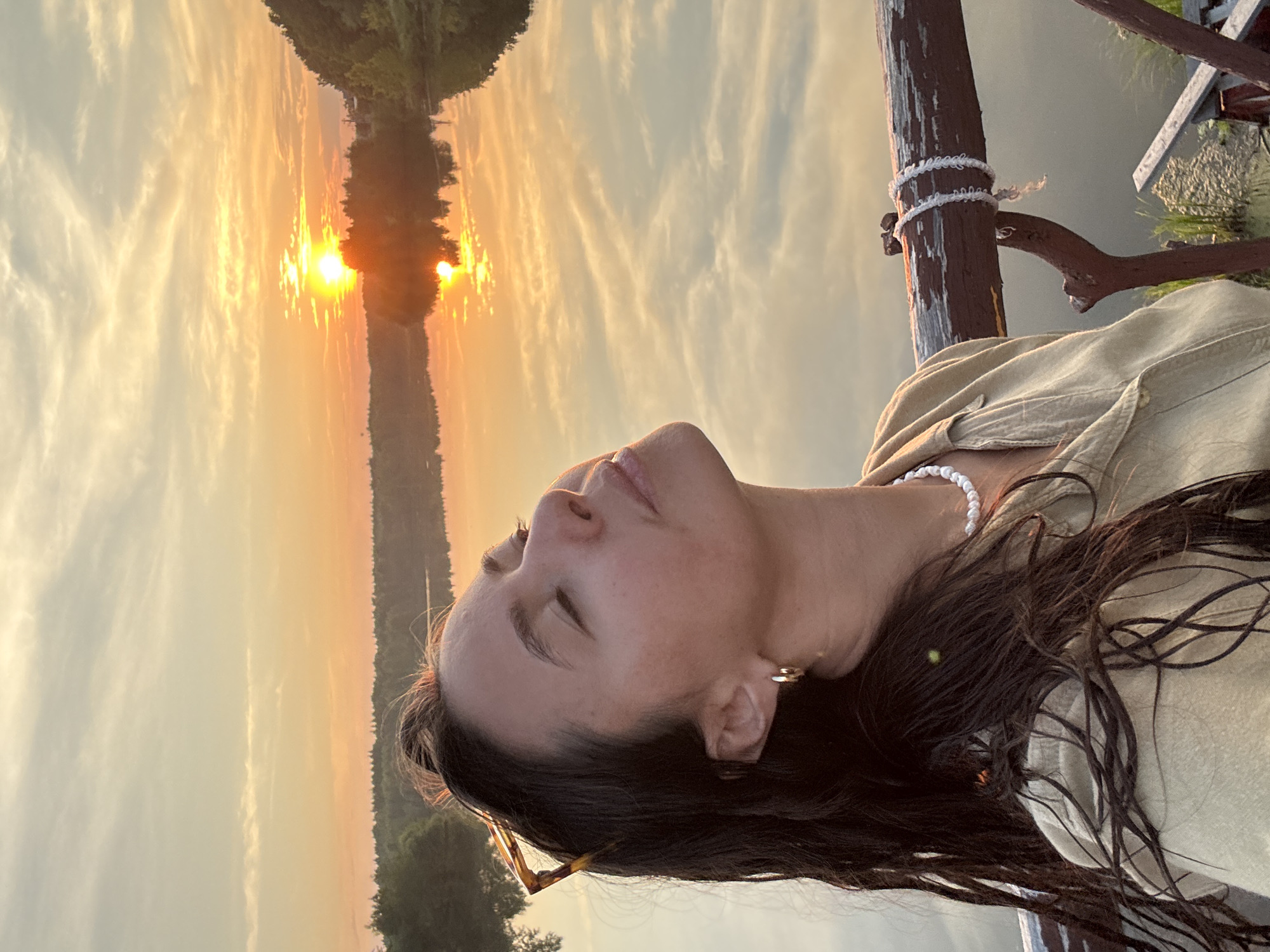 A medium shot captures a woman with dark, wet hair and closed eyes, her face tilted upwards towards a sunset over a body of water. She is wearing a light-colored collared shirt and a pearl necklace. Her head is angled so that her profile is visible, with sunglasses perched on top of her head. The background features a vibrant sunset with orange and yellow hues, reflected in the calm water. Trees and foliage line the opposite bank of the water. The woman appears to be standing on a wooden structure, possibly a bridge or pier, with a weathered railing in the foreground. The image conveys a sense of peace and tranquility. The location is identified as Kamysh, Russia, suggesting a rural or natural setting within the city.