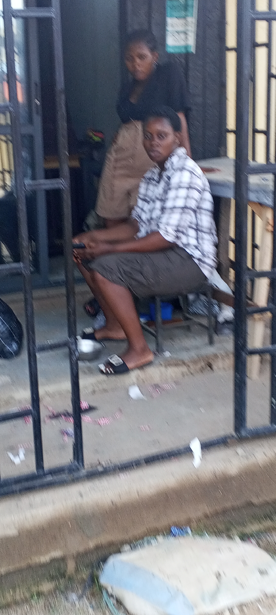 Two people are visible through a black iron barred fence, likely in Sabon Tasha, Nigeria. The person in the foreground is sitting on a small stool, wearing a plaid shirt and dark shorts, and appears to be using a mobile device. They are wearing sandals. The other person is standing in the background, wearing a dark top and light-colored skirt. There is trash and debris on the ground near the seated individual.