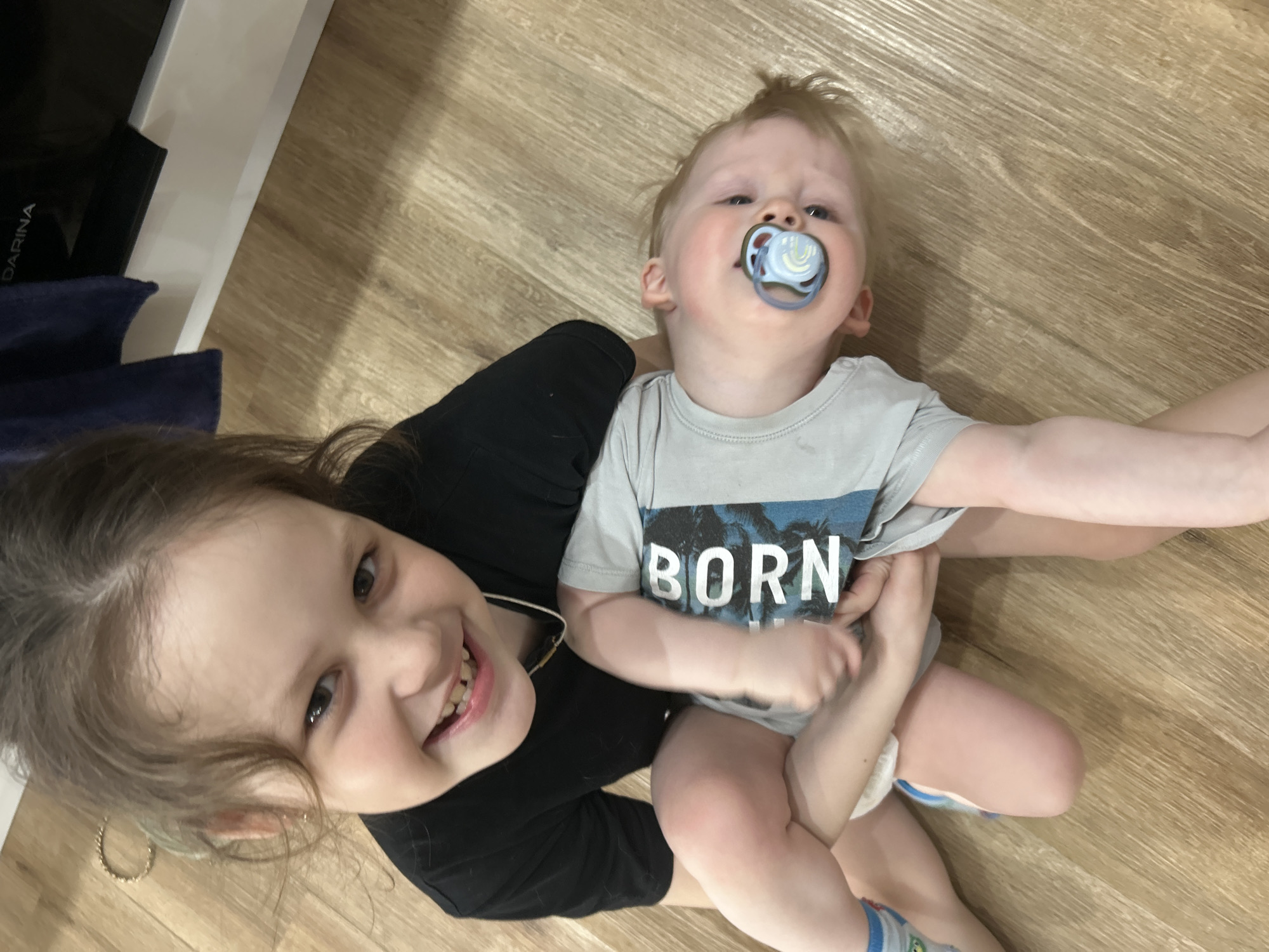 A young girl with brown hair smiles at the camera while holding a baby. The baby has blond hair and a pacifier in its mouth. The baby is wearing a gray shirt with the word "BORN" printed on it. They are sitting on a wooden floor. There are clothes next to them on the floor. It is not possible to verify the location of the city of Chaikovsky, Russia, in the scene.