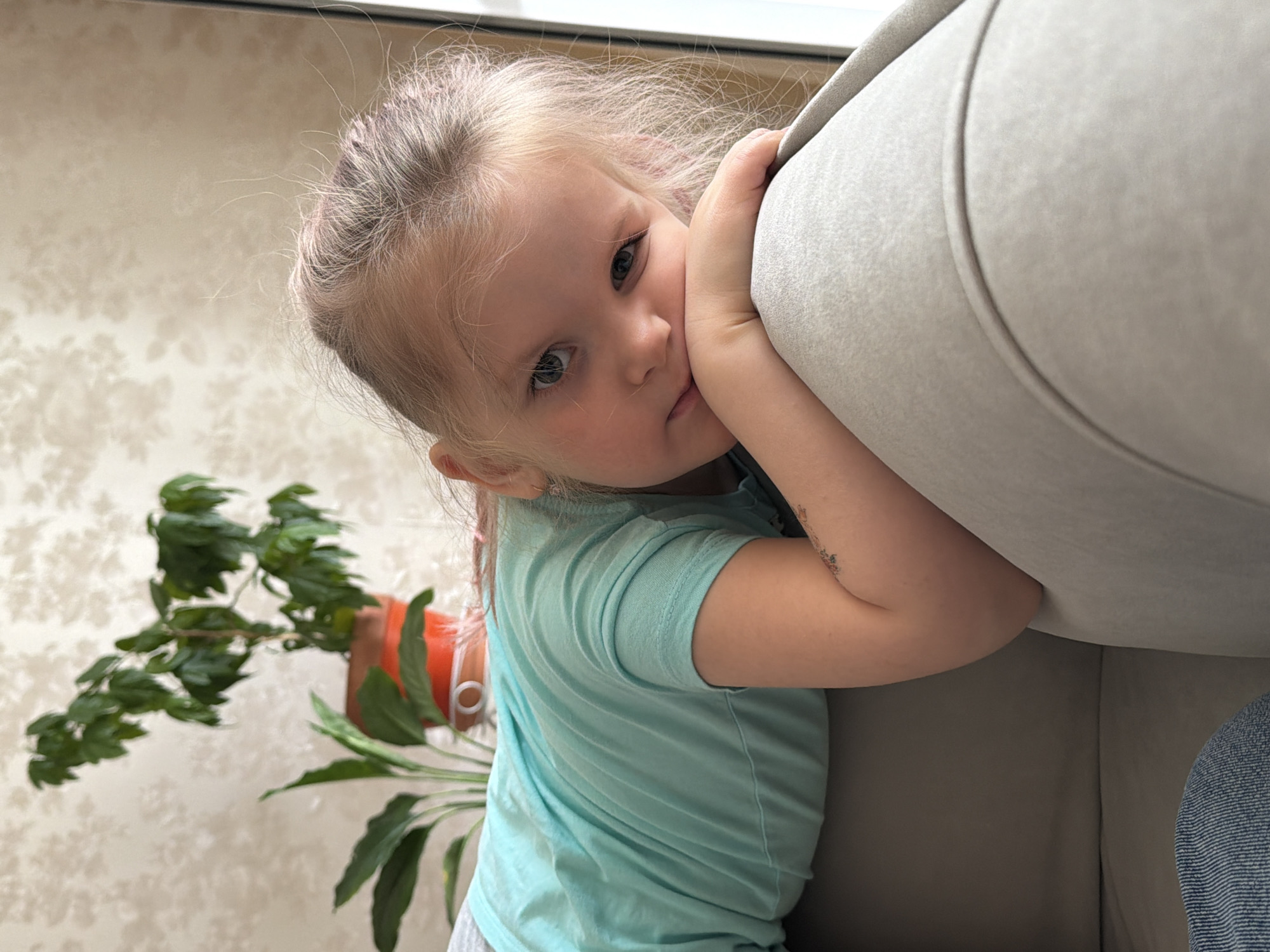 A young child with light-colored hair, pulled back, looks directly at the camera with large, dark eyes. She is leaning against a light-colored, upholstered sofa or armchair, wrapping her arms around the furniture and resting her face partially against it. Her expression appears innocent and somewhat contemplative or shy.
The child is wearing a light blue or teal short-sleeved t-shirt. A faint dark mark or design is visible on her right upper arm, possibly a temporary drawing. In the background, a light-colored wall features a subtle patterned wallpaper. To the left, a green houseplant in an orange pot is visible. Bright, natural light enters from the top of the frame, indicating daytime. A glimpse of blue textured fabric, possibly denim, is seen in the lower right corner. The scene is indoors, likely a residential setting in Chaikovsky, Russia. There is no visible text.
