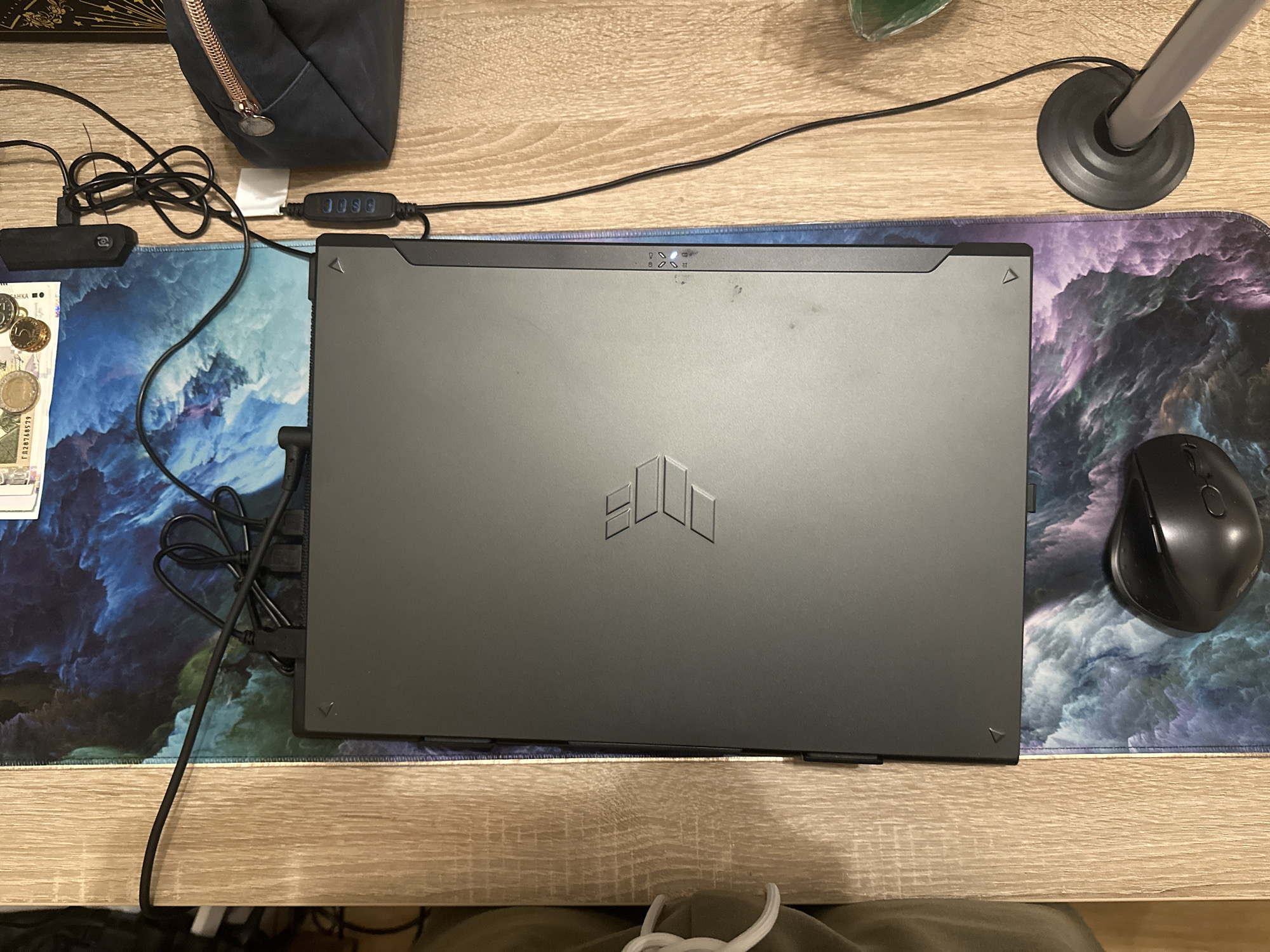 The scene depicts a top-down view of a workstation or desk. The location cannot be determined from the image. A closed, dark grey or black ASUS TUF Gaming series laptop, identifiable by its stylized debossed logo, is centrally positioned on a large mouse pad. The mouse pad features an abstract pattern resembling blue, purple, and white clouds or nebulae. The underlying surface is a light-colored wooden desk with a visible grain.
Multiple black cables emerge from the rear of the laptop; some are bundled together. One cable connects to a small, black rectangular device, likely a USB hub, positioned on the mouse pad in the upper left area. Another cable extends from the right side of the laptop to an inline black control unit featuring four circular buttons, typically used for adjusting an LED light source.
To the upper left, on the mouse pad, scattered currency is visible, including several coins and a 100 Bulgarian Lev banknote (serial number ГД28768579 is partially visible). In the upper right quadrant of the wooden desk, a dark blue or black fabric pouch or small bag with a prominent rose gold or copper zipper is present.
A black ergonomic computer mouse rests on the mouse pad in the lower left section. In the lower right corner, on the wooden desk surface, the circular black base of a desk lamp or similar stand is visible. The right edge of the laptop reveals three illuminated indicator lights (two white, one amber) with associated icons, likely denoting power, battery status, and disk activity. A light-colored fabric object is partially visible on the far left, and a translucent green object is partially visible on the far right.