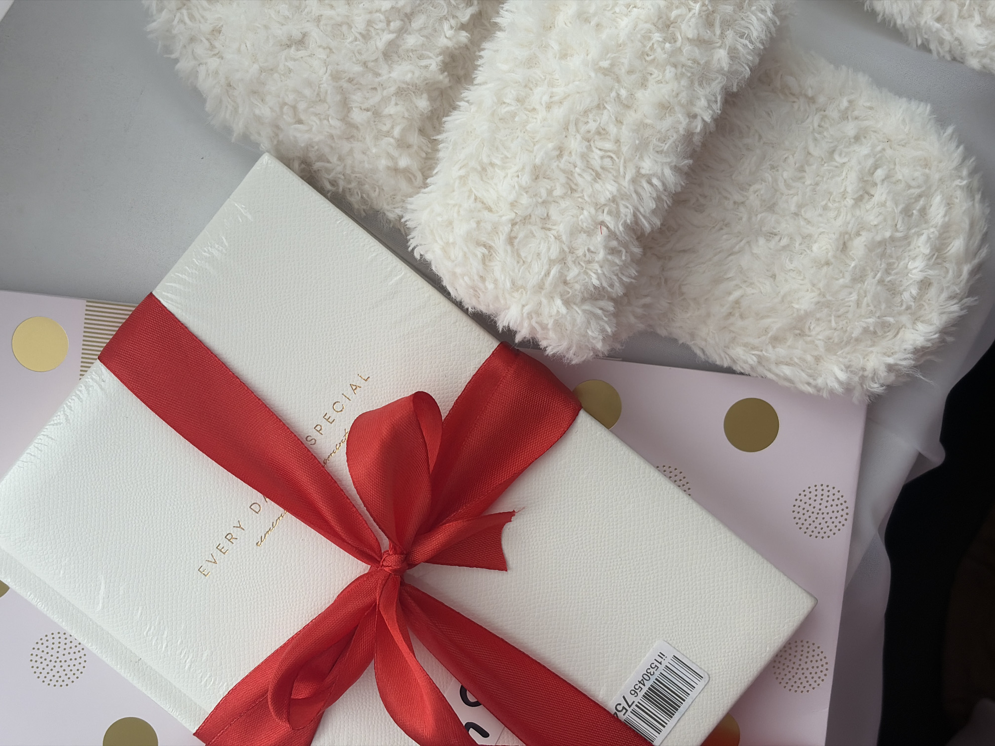 The image shows a gift-giving scene, featuring wrapped presents and soft, white material. A white, rectangular box wrapped with a red ribbon is prominently placed in the center. The box has "EVERY DAY SPECIAL" printed on it. It sits atop another gift box with pink and gold polka dot wrapping paper. A soft, fluffy white material rests partially on top of the boxes and extends toward the upper right corner of the frame. The objects rest on a white surface. The setting appears indoors, with diffused lighting. There's no immediate indication of a specific location like Saint Petersburg, Russia.